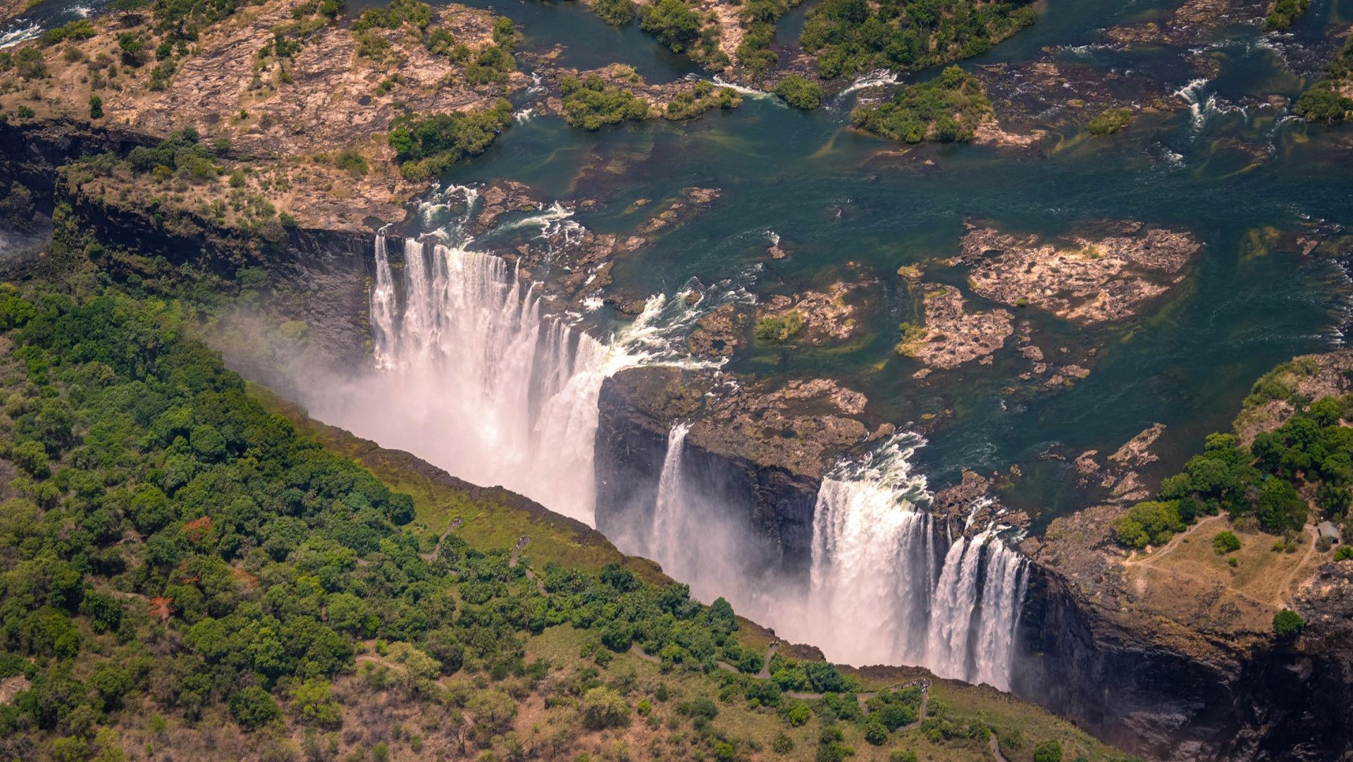 Waterfall cascading over a cliff edge, creating mist; green and white water, gray sky.