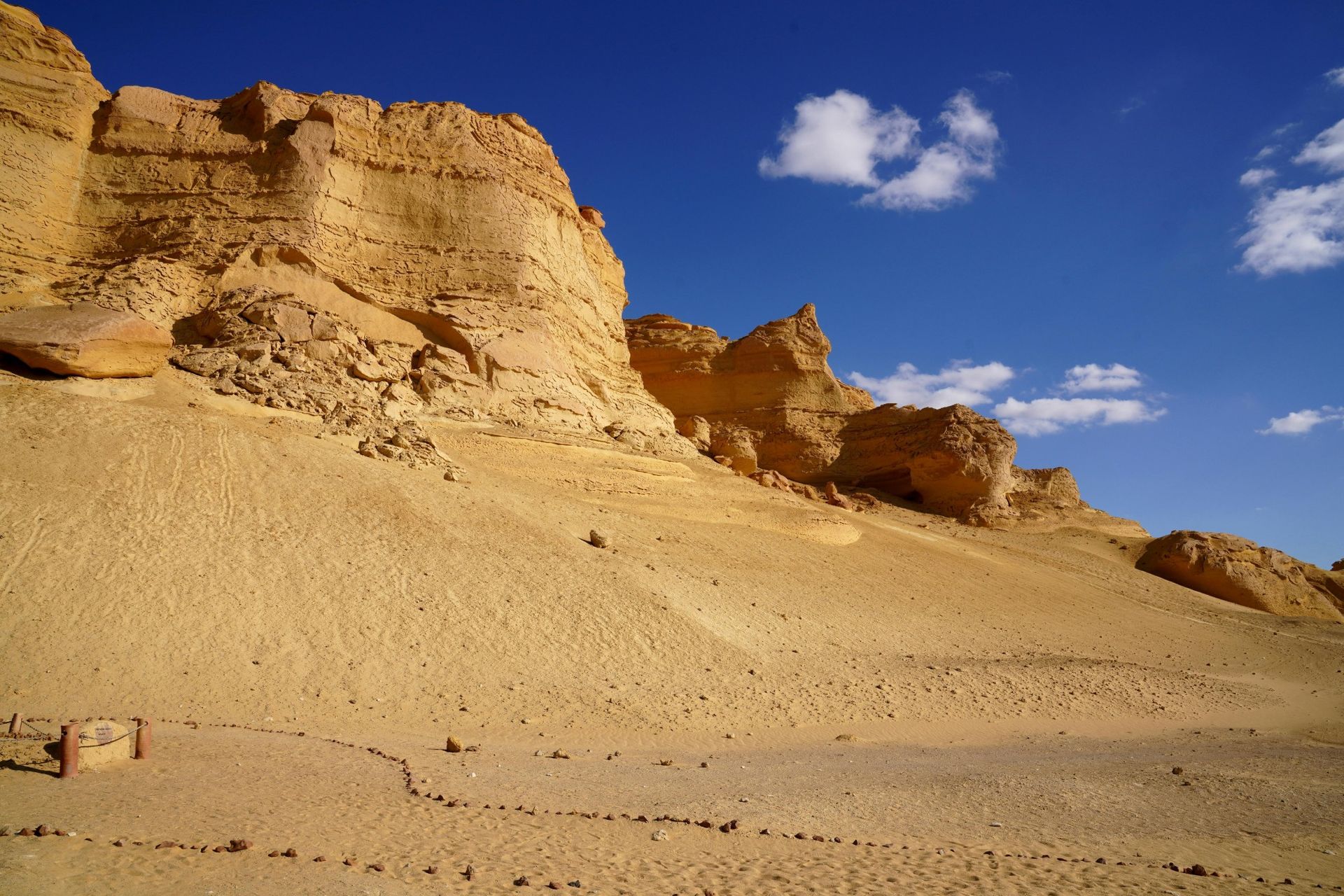 Desert landscape with layered tan cliffs under a bright blue sky with a few clouds.