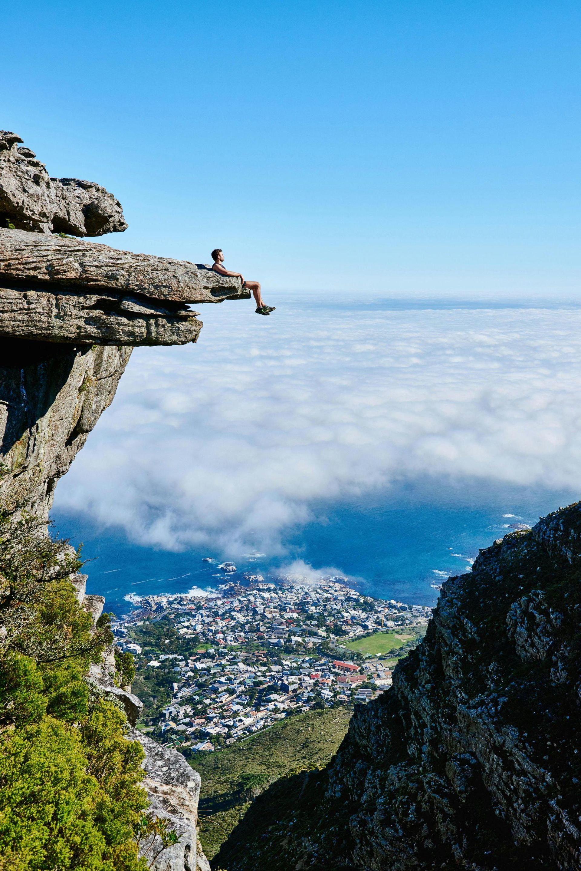 Person sitting on a rock ledge, looking out over a city, blue sky, and clouds.