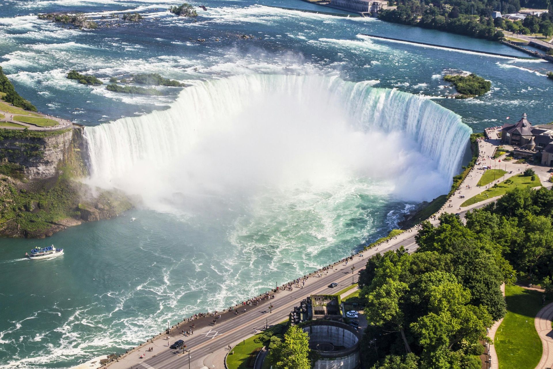 Niagara Falls, a large waterfall with a foamy base, viewed from above, surrounded by green vegetation and buildings.