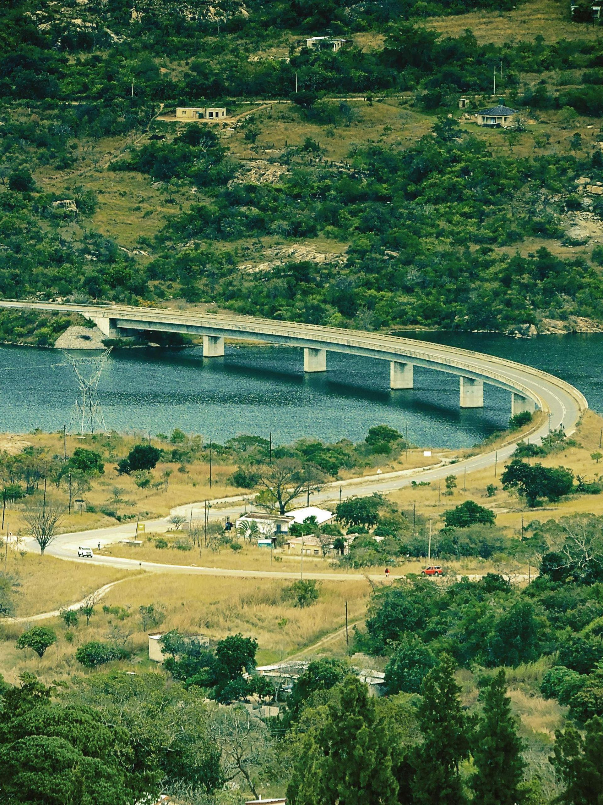 Curving bridge over blue water, surrounded by green trees and dry, grassy land.