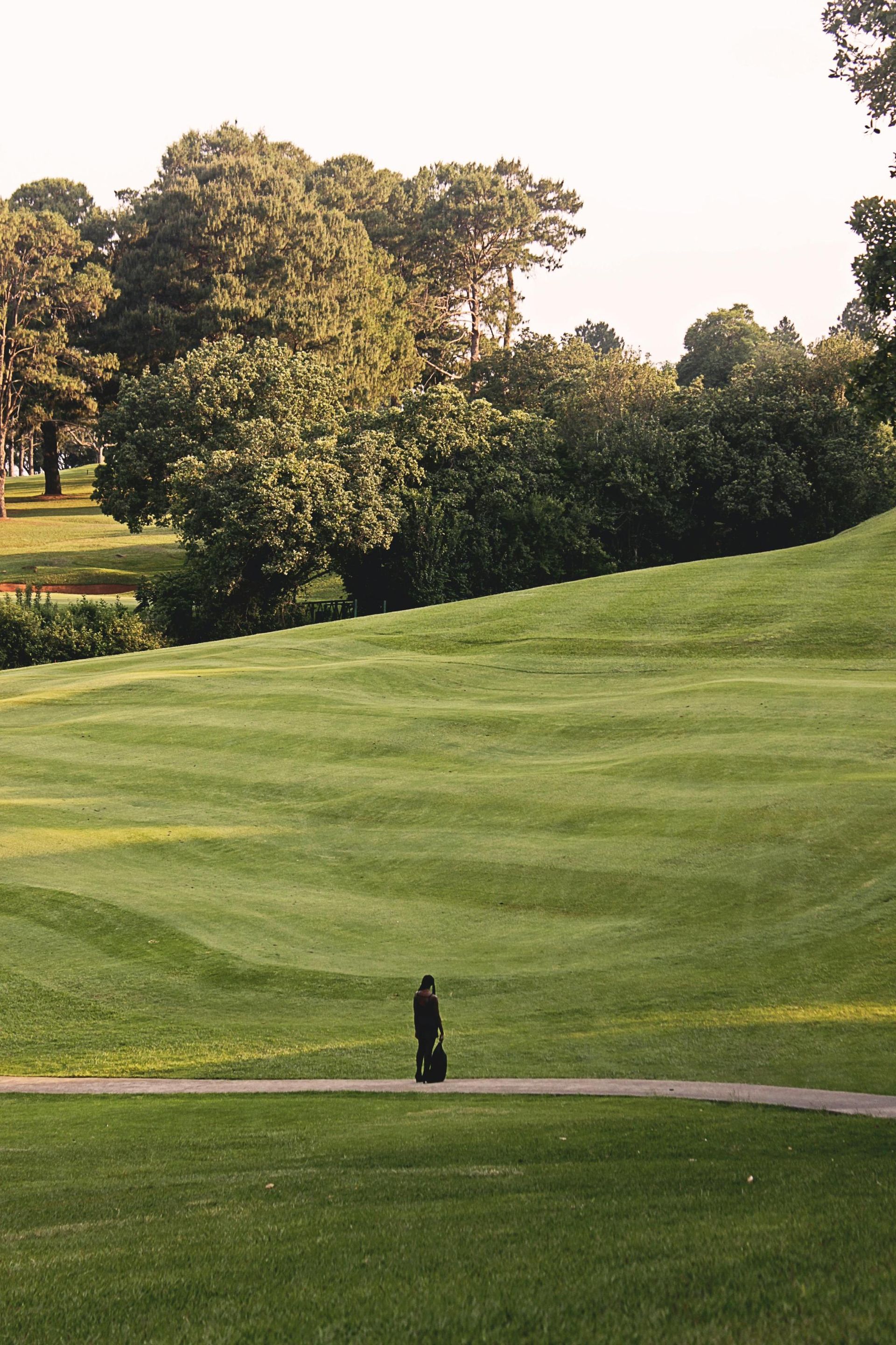 Person walking on a golf course path, carrying a golf bag. Green grass and trees fill the sunny landscape.