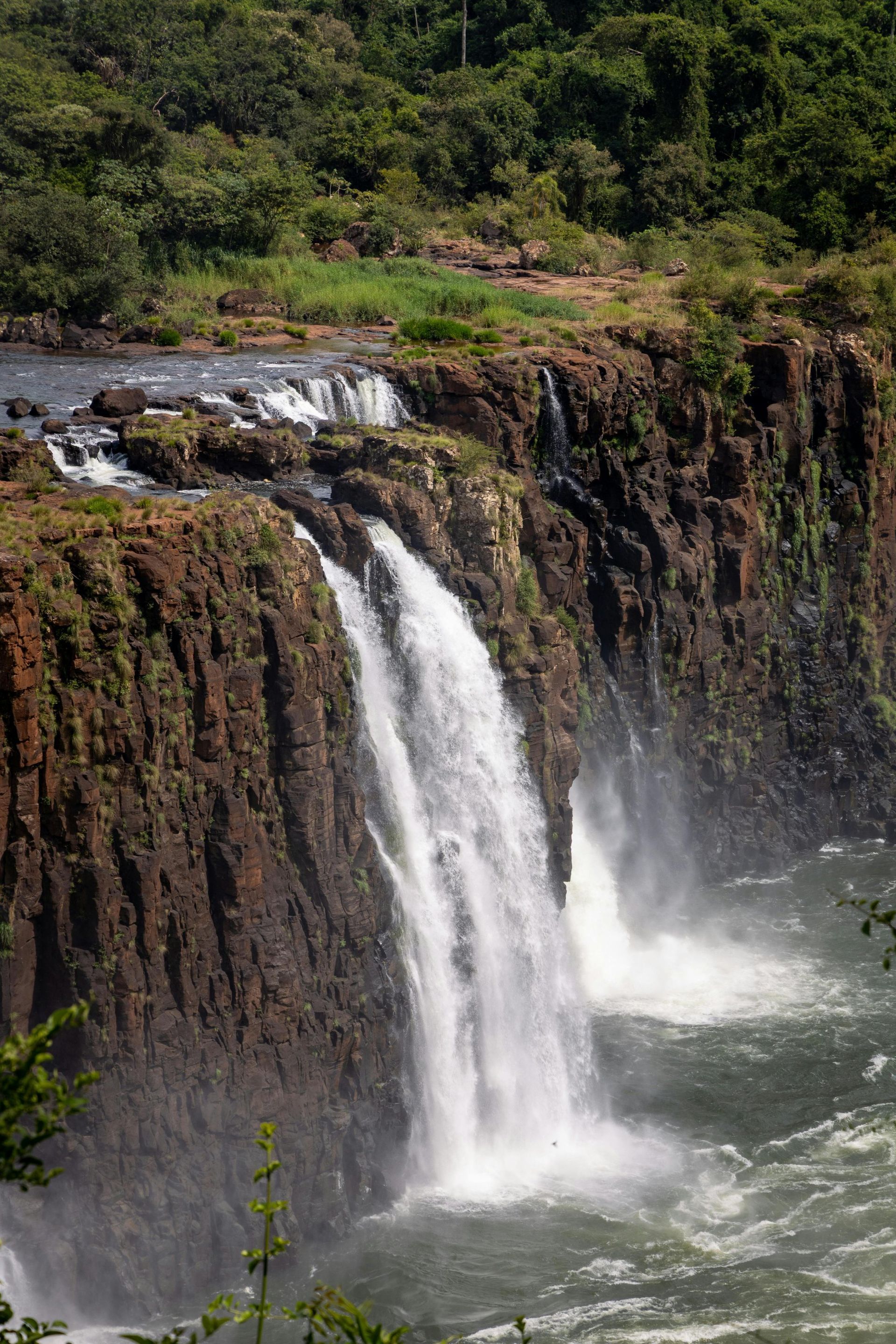 Waterfalls cascading over rocky cliffs into a turbulent river. Lush green forest in the background.