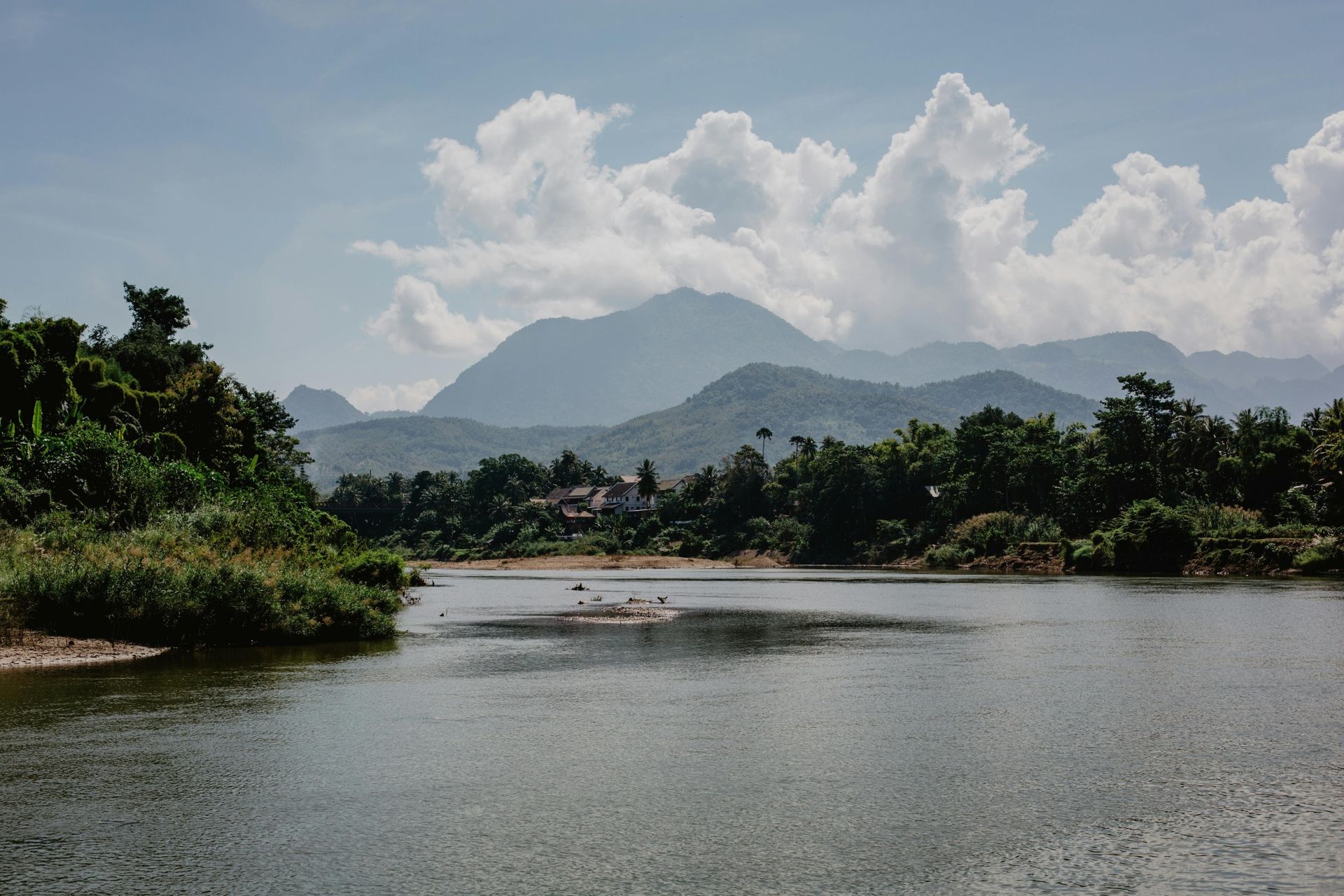 River with mountains and cloudy sky, surrounded by green trees.