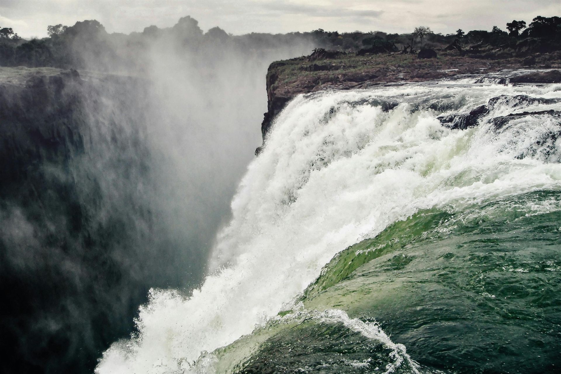 Waterfall cascading over a cliff, creating mist; green vegetation at the edge, overcast sky.
