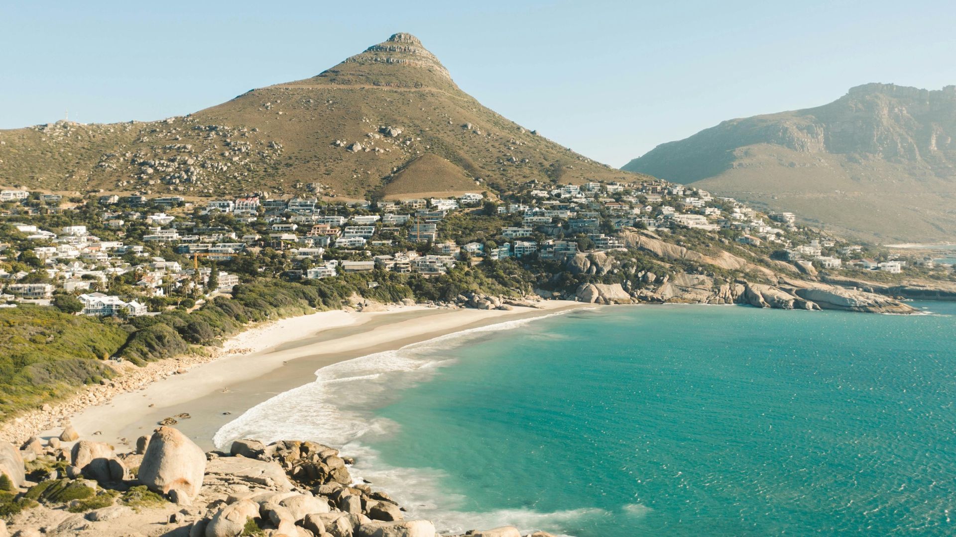 Beach with turquoise water, white sand, and houses on a hill, dominated by a mountain.