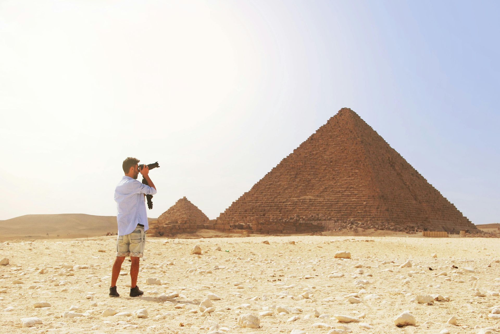 Man taking a photo of the Great Pyramids in Egypt, standing on sand under a sunny sky.