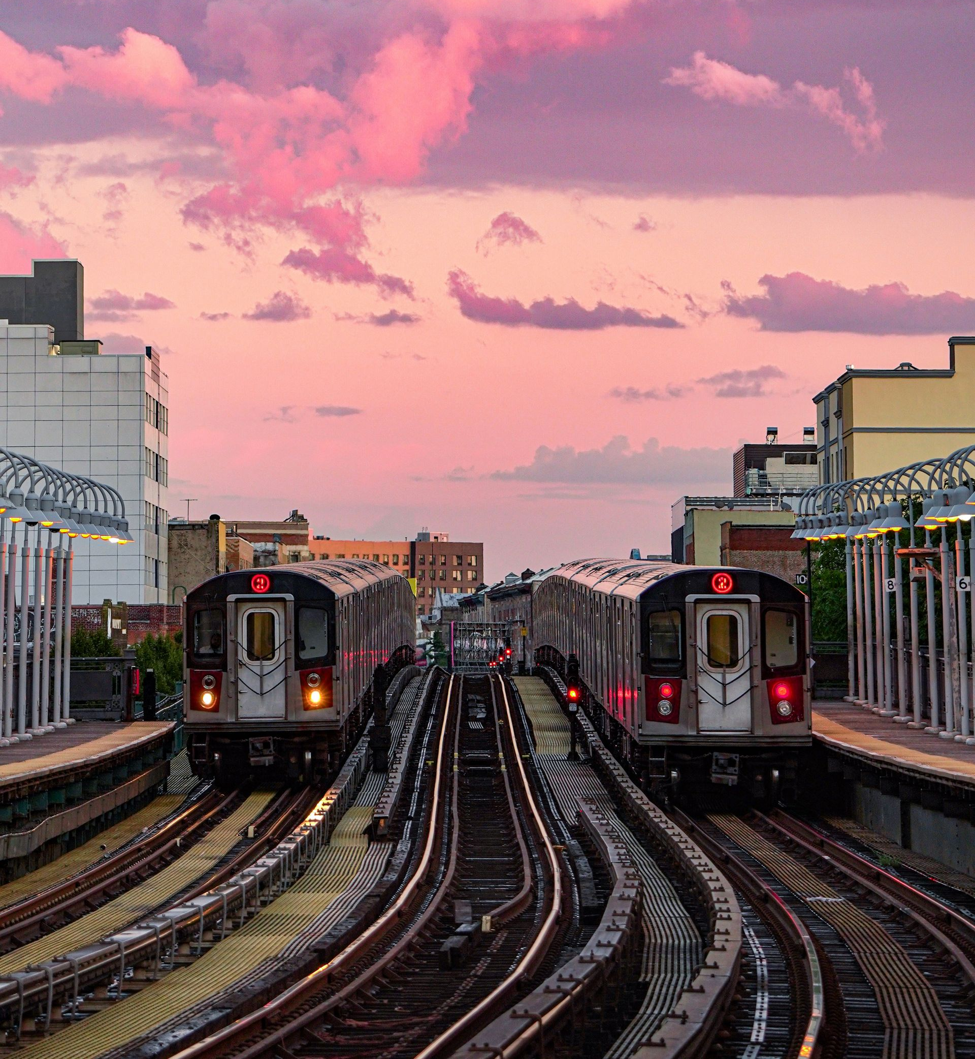 Two subway trains at a station, tracks curving into the distance under a pink sunset sky.