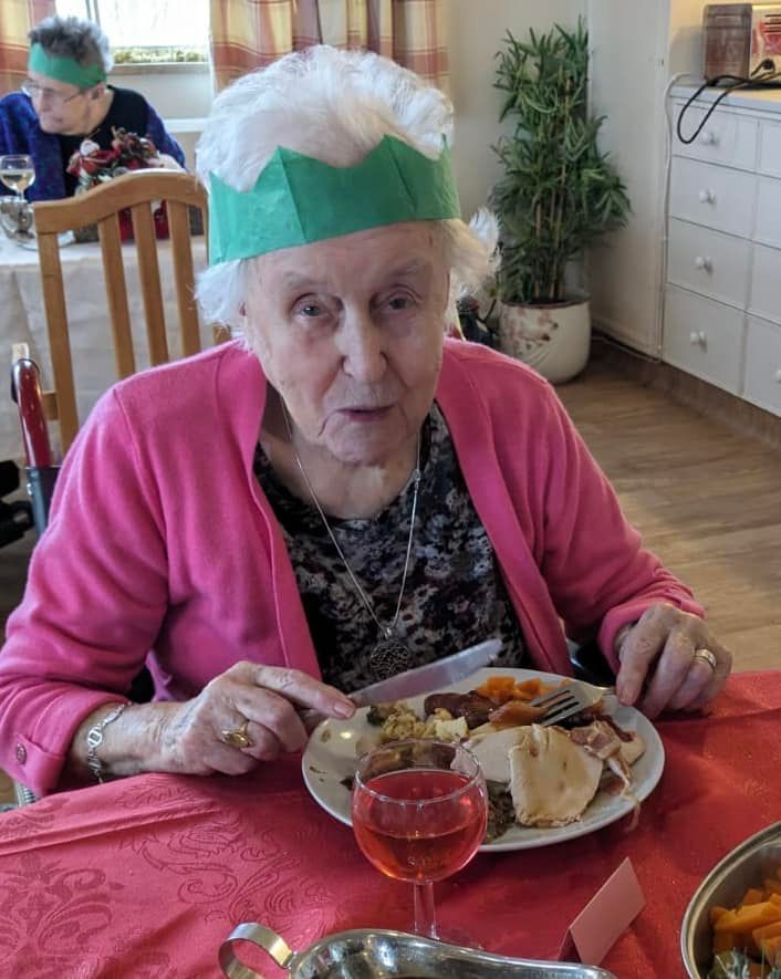An elderly woman is sitting at a table with a plate of food and a glass of wine.