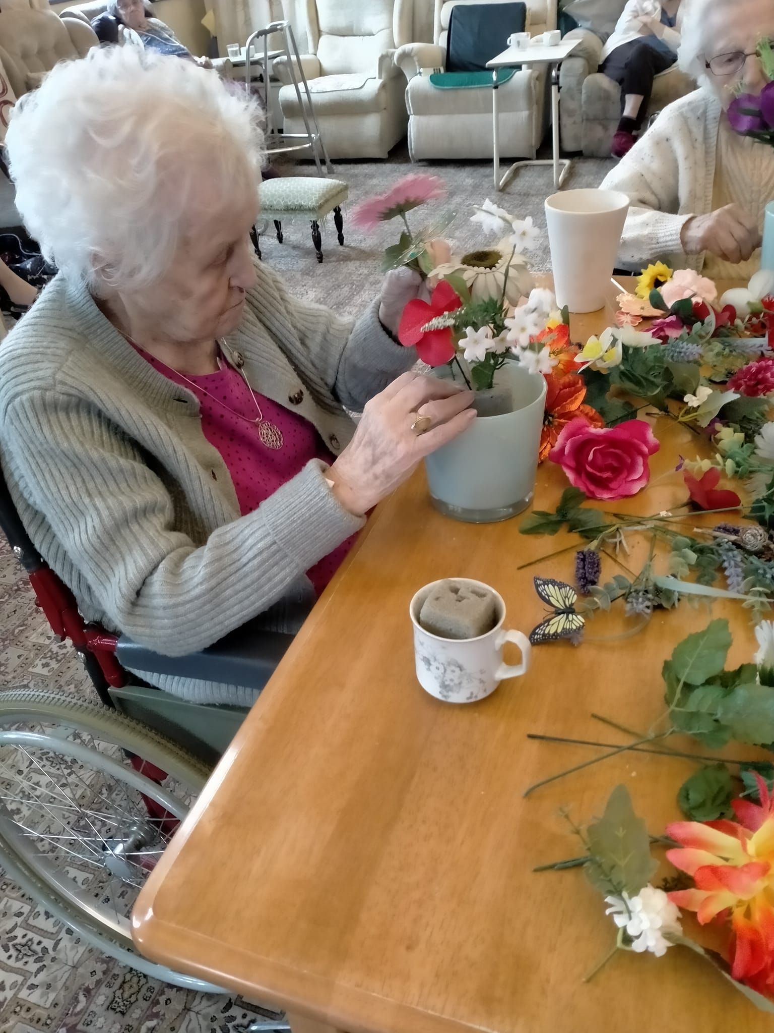 A woman in a wheelchair is sitting at a table with flowers and a cup of coffee.