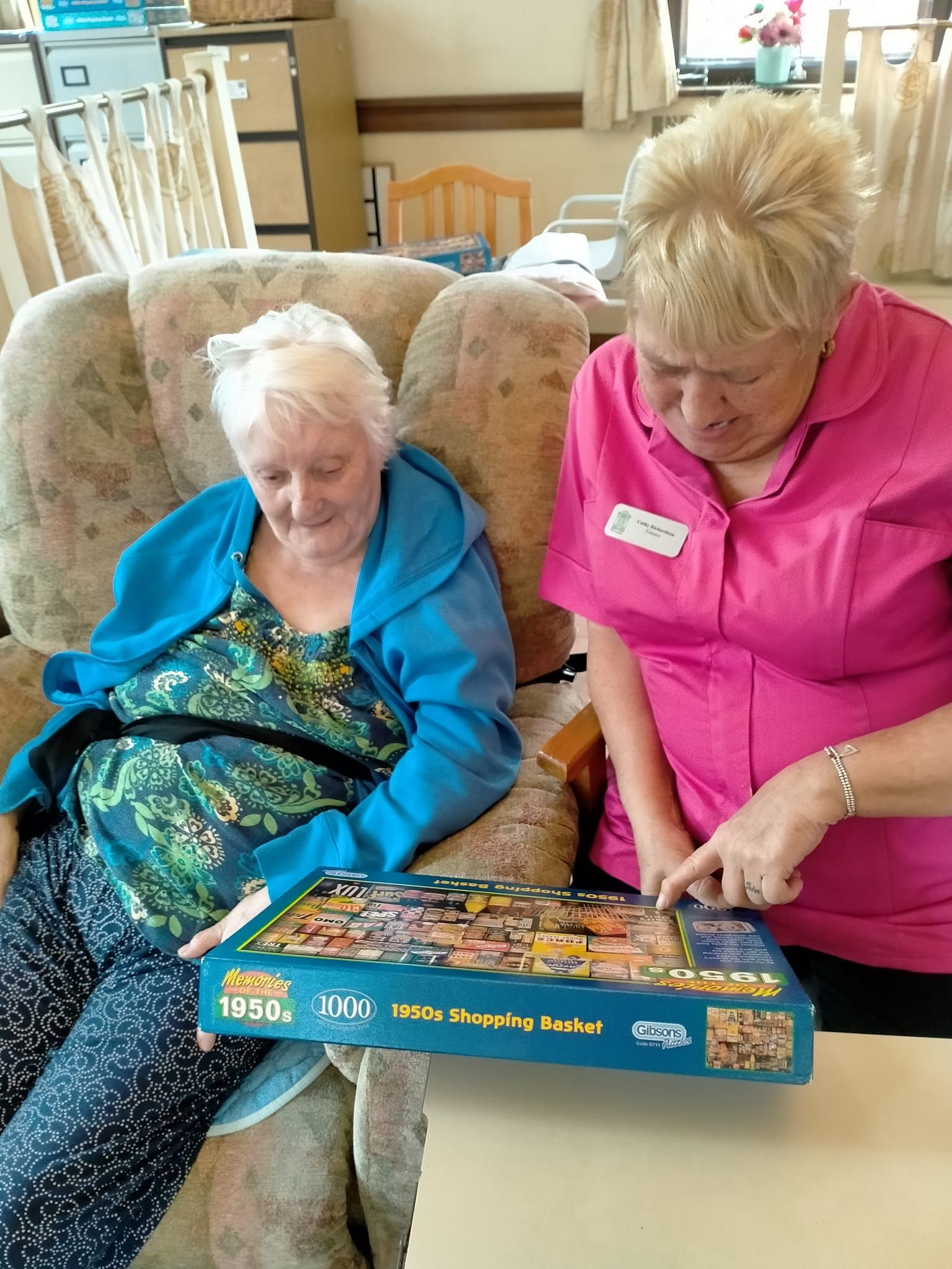 A woman is sitting in a chair next to another woman holding a puzzle.