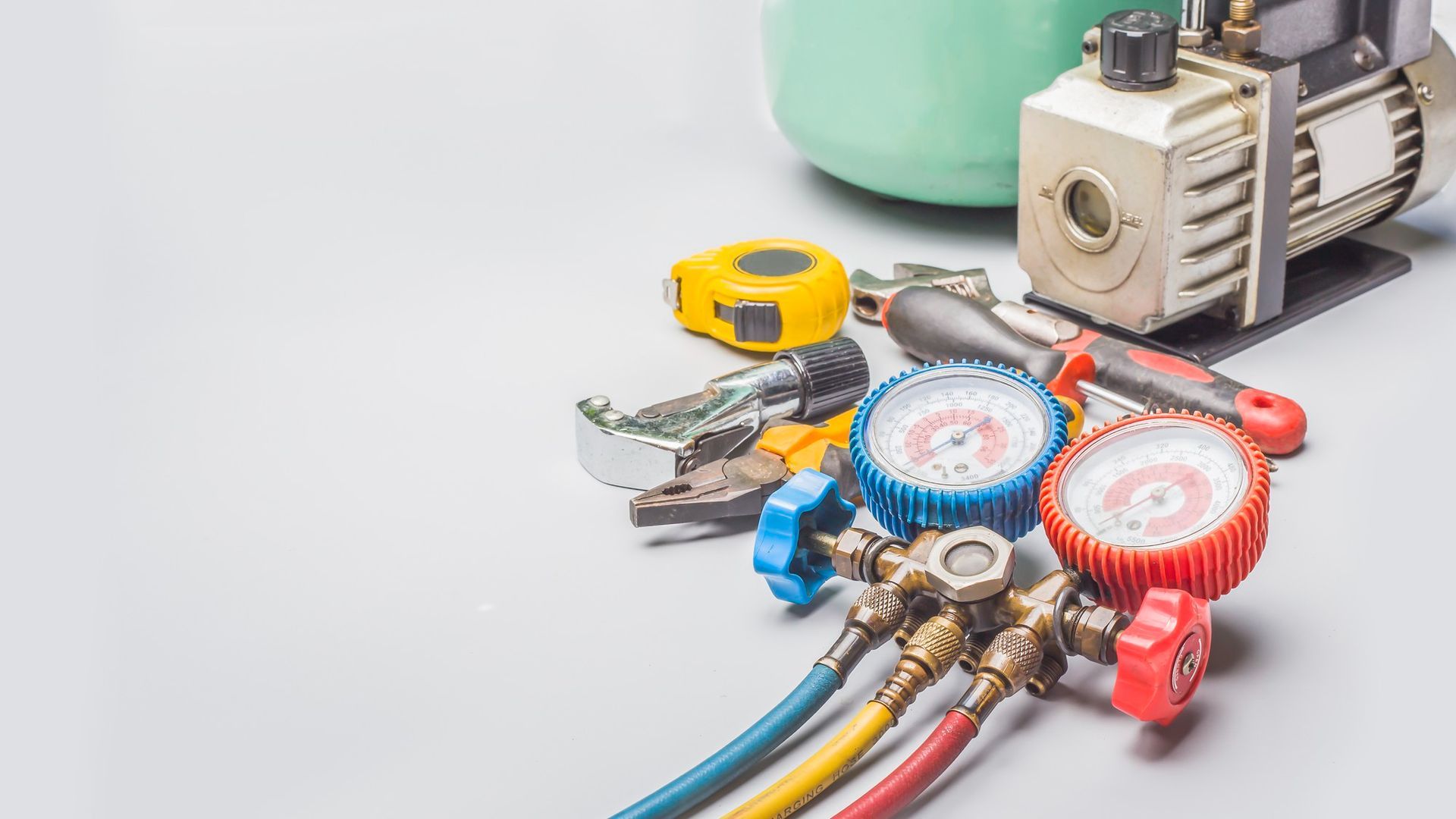 HVAC repair tools including gauges, pump, refrigerant tank, and wrenches on a light surface.