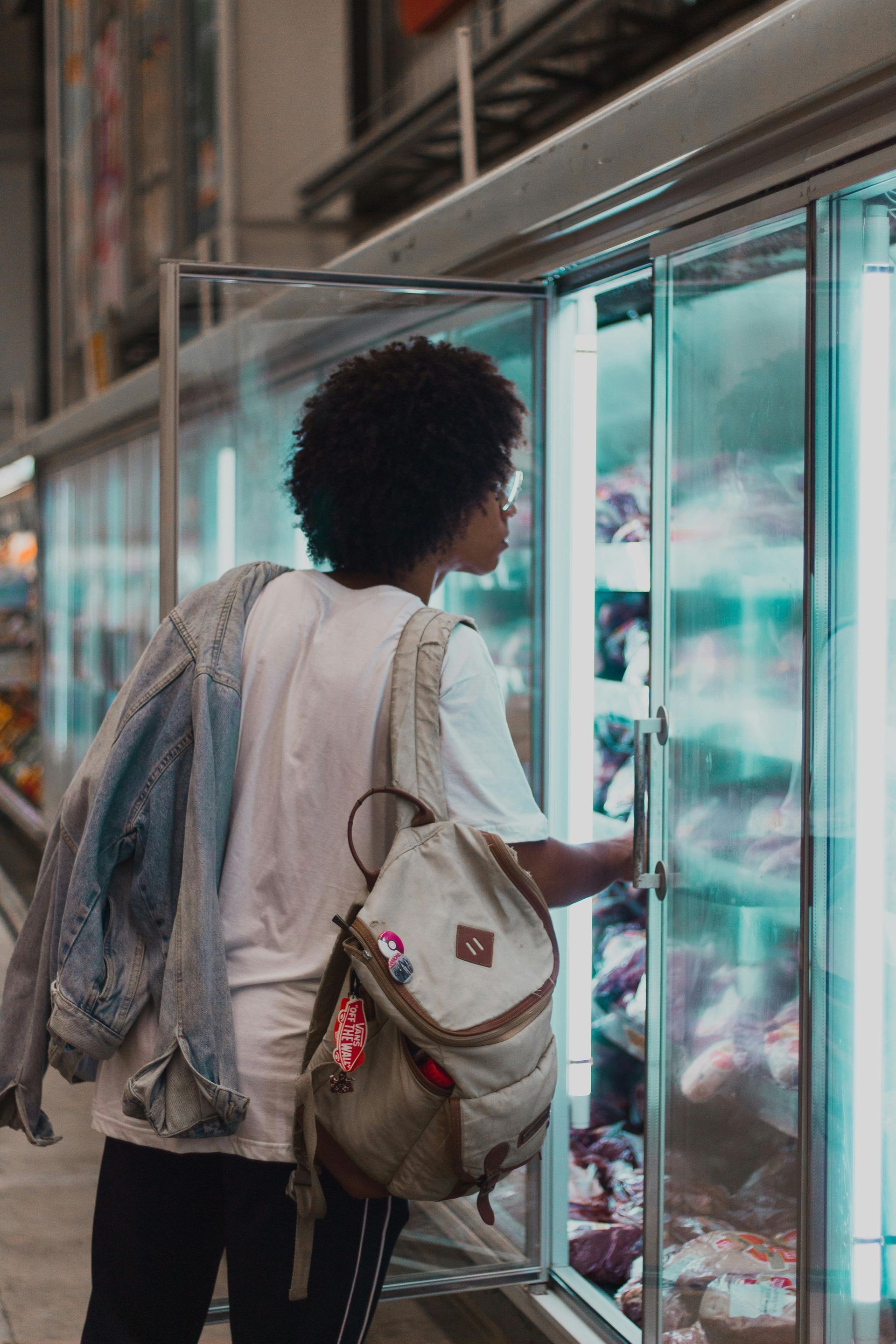 Person reaching into a lit freezer in a grocery store, carrying a backpack and denim jacket.
