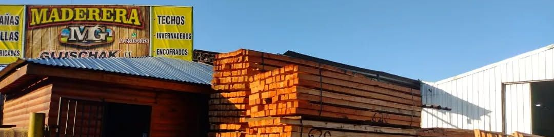 Un almacén de madera con tablones de madera apilados, edificios y un cartel bajo un cielo azul.