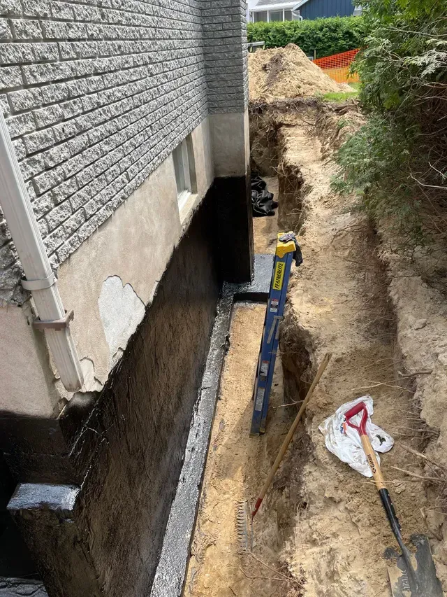 Un chantier de réparation de fondations avec une tranchée creusée le long d'un mur de maison, montrant une base en béton imperméabilisé et en gravier à nu.