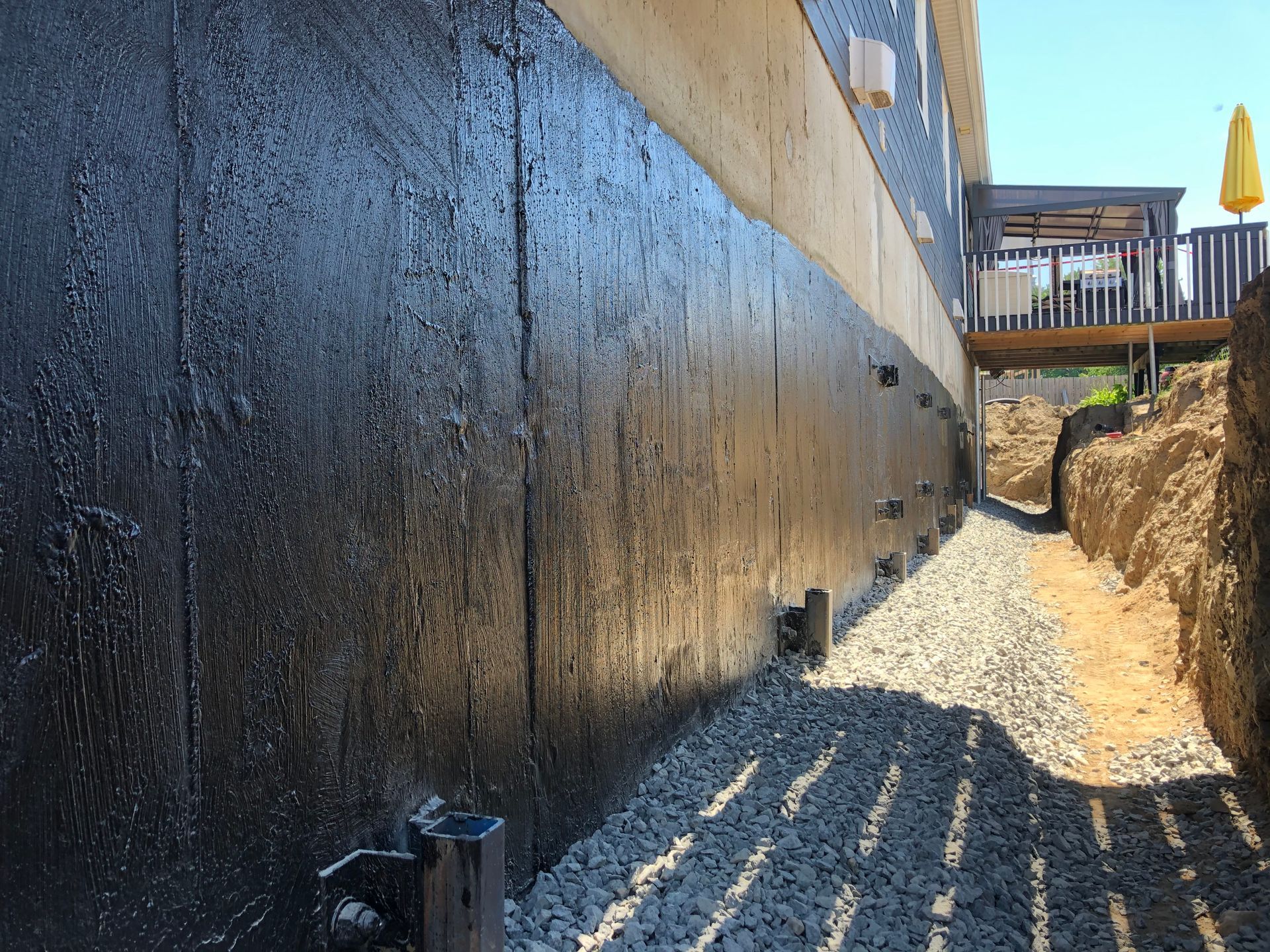 Un mur de fondation résidentiel recouvert d'un enduit d'étanchéité noir, avec une tranchée excavée remplie de gravier à côté.