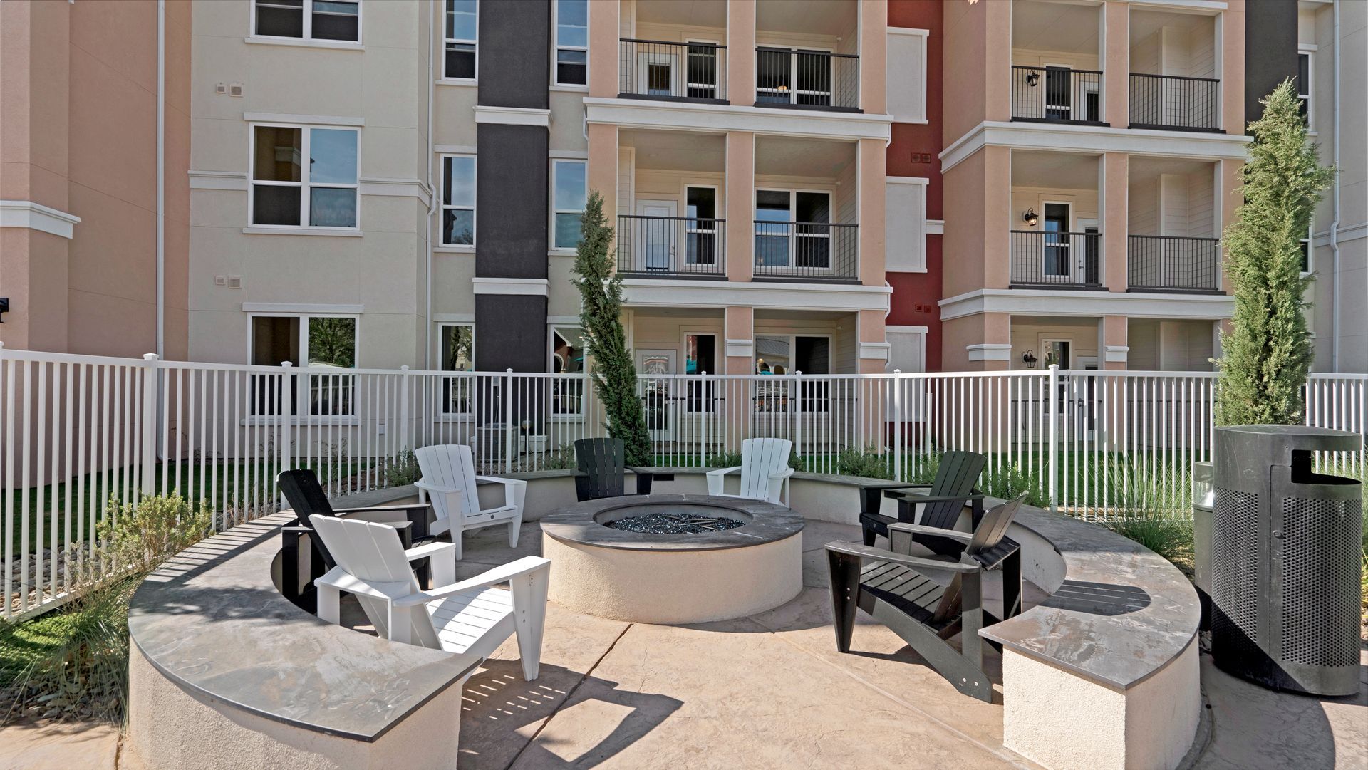 A fire pit is surrounded by chairs and a fence in front of a building.