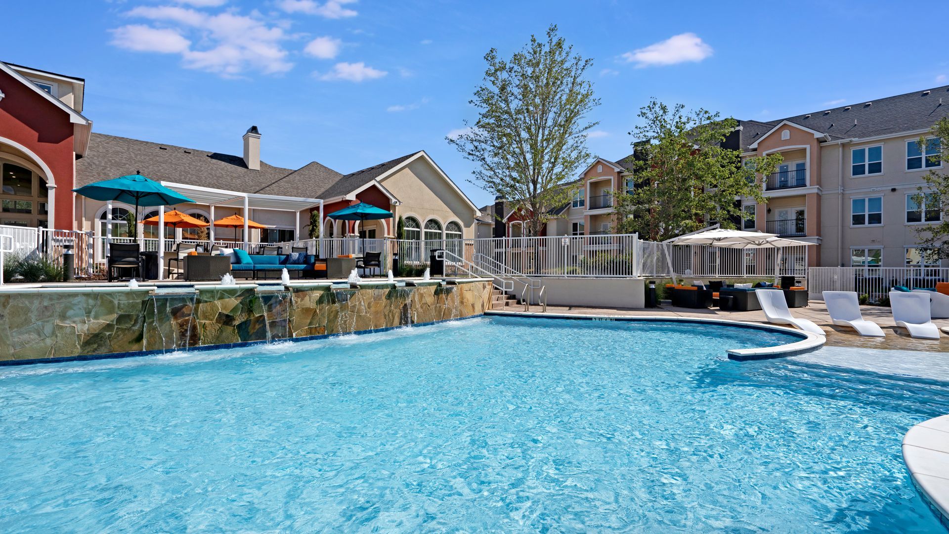 A large swimming pool surrounded by chairs and umbrellas in front of a building.