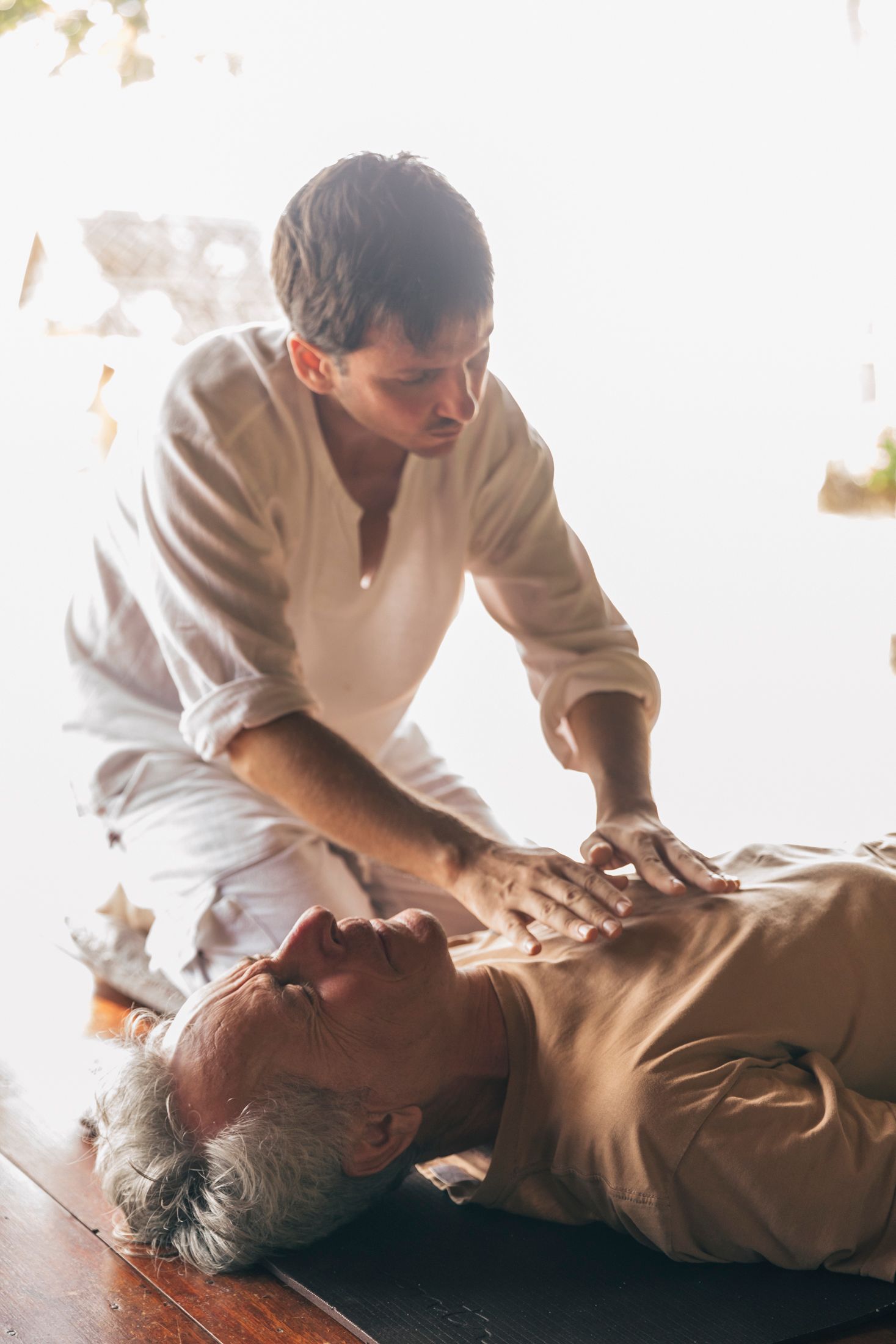 Man performing chest compressions on an older man lying down; both are in white clothing.