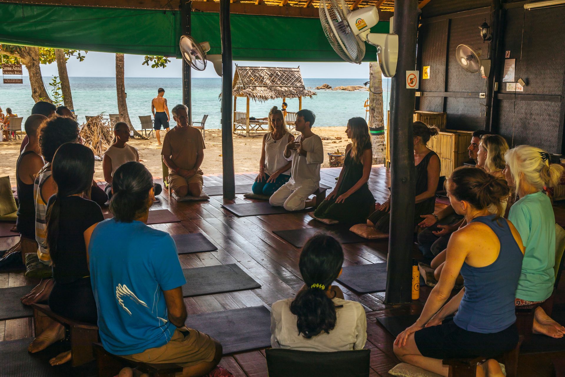 A group of people sit in a circle on a wooden floor near a beach, participating in a workshop or class.