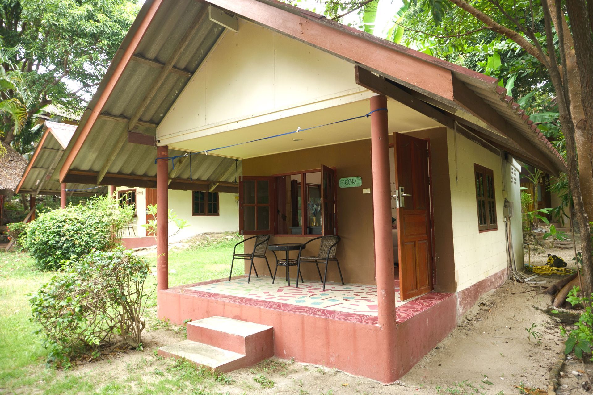 Bungalow with brown roof and trim, beige walls, small porch with table and chairs, set amidst green foliage.