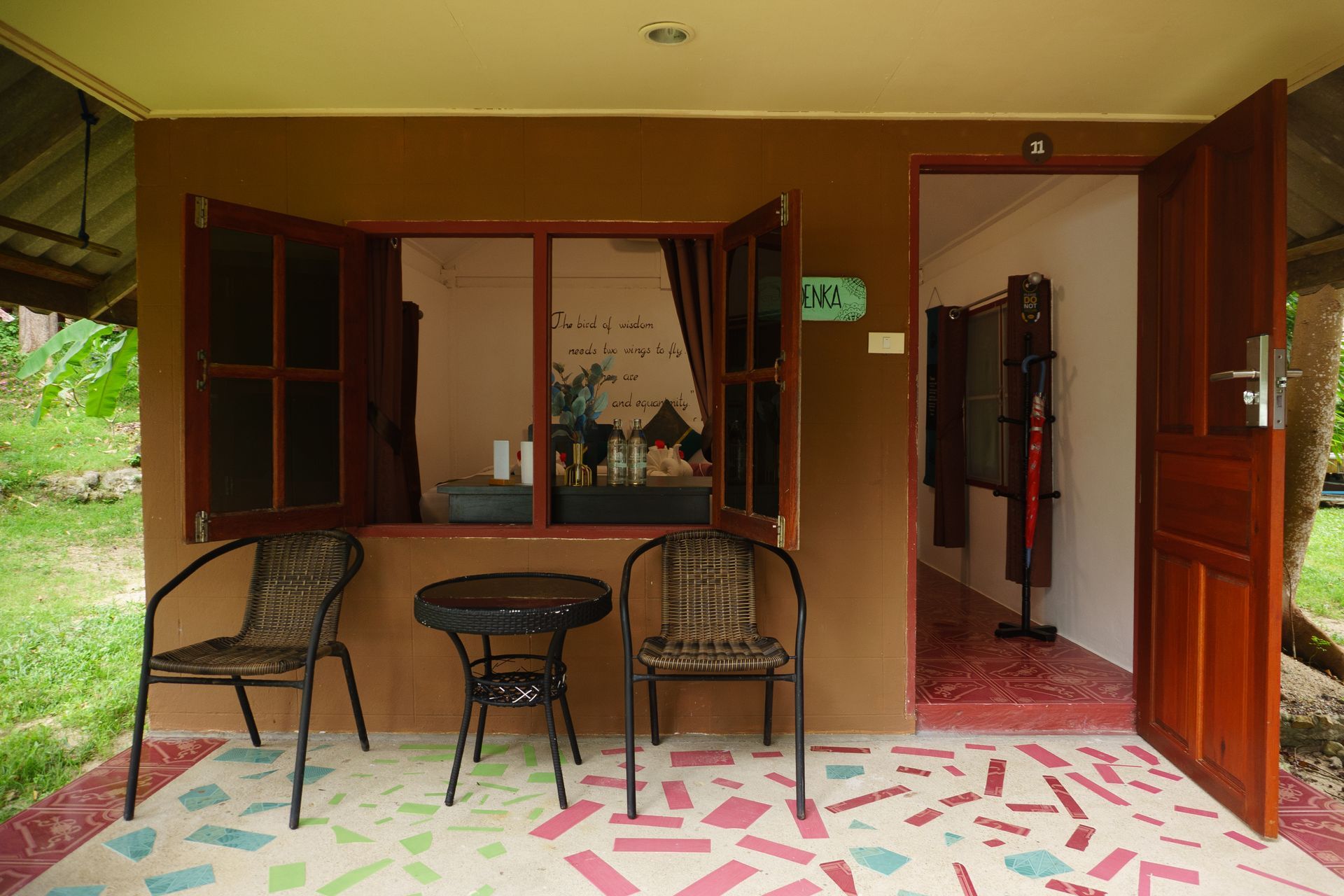 Brown building exterior with open door and window, two chairs, and a small table on a mosaic-tiled porch.