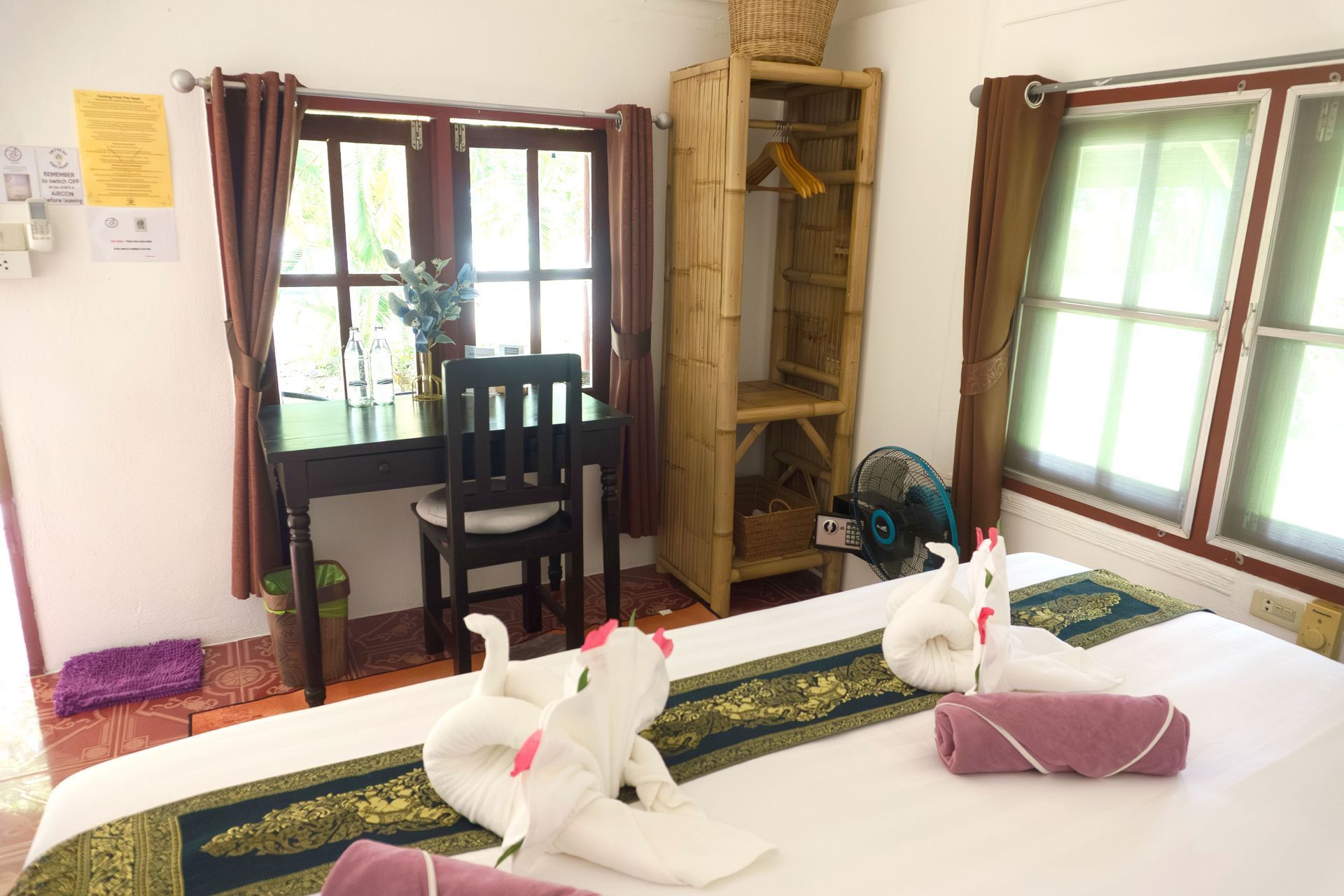 Bedroom with bed, desk, bamboo closet, windows with brown curtains, and towels shaped like swans.