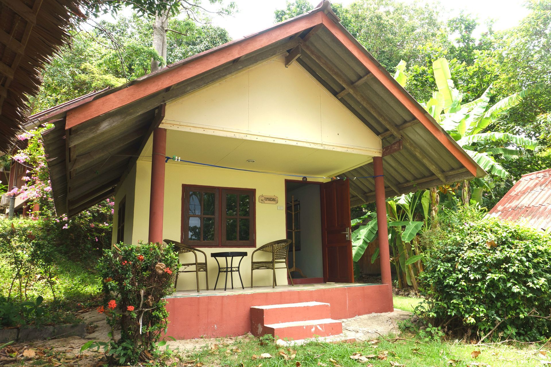 Tan bungalow with red trim and a porch, set amongst tropical foliage.