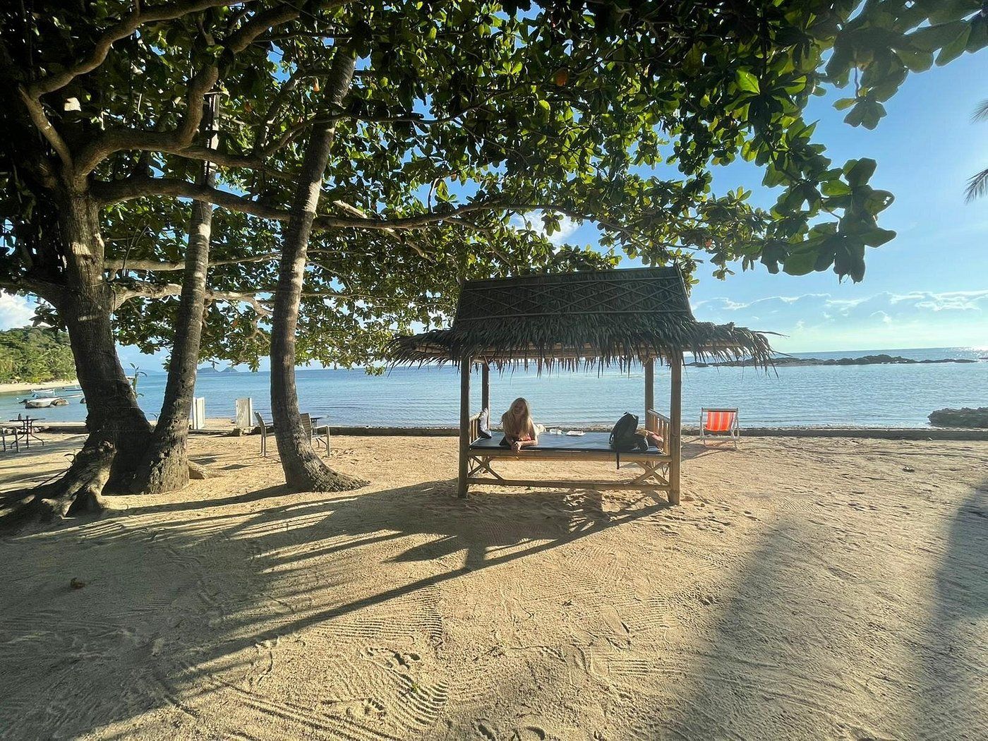Beach scene with a hut, trees, and people. Sunlight, water, and blue sky.