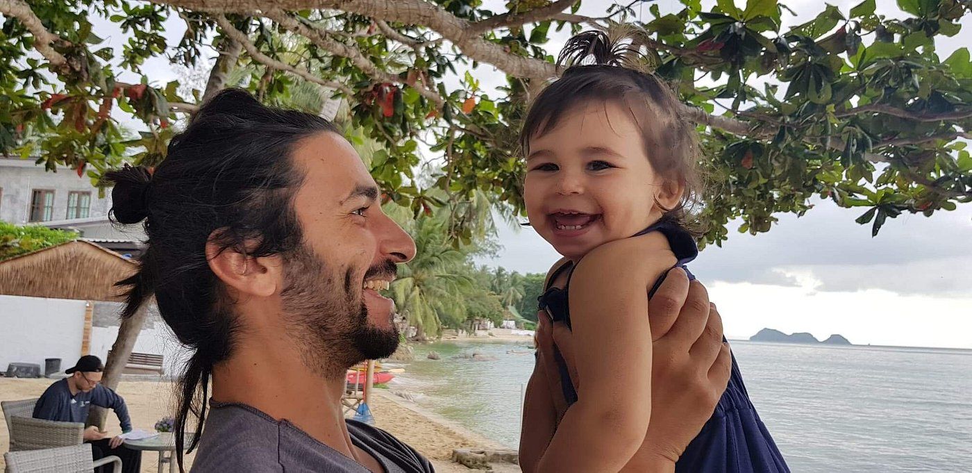 Man holding a smiling baby near a beach; they're under tree branches. Man has dark hair, beard. Baby wears a blue dress.