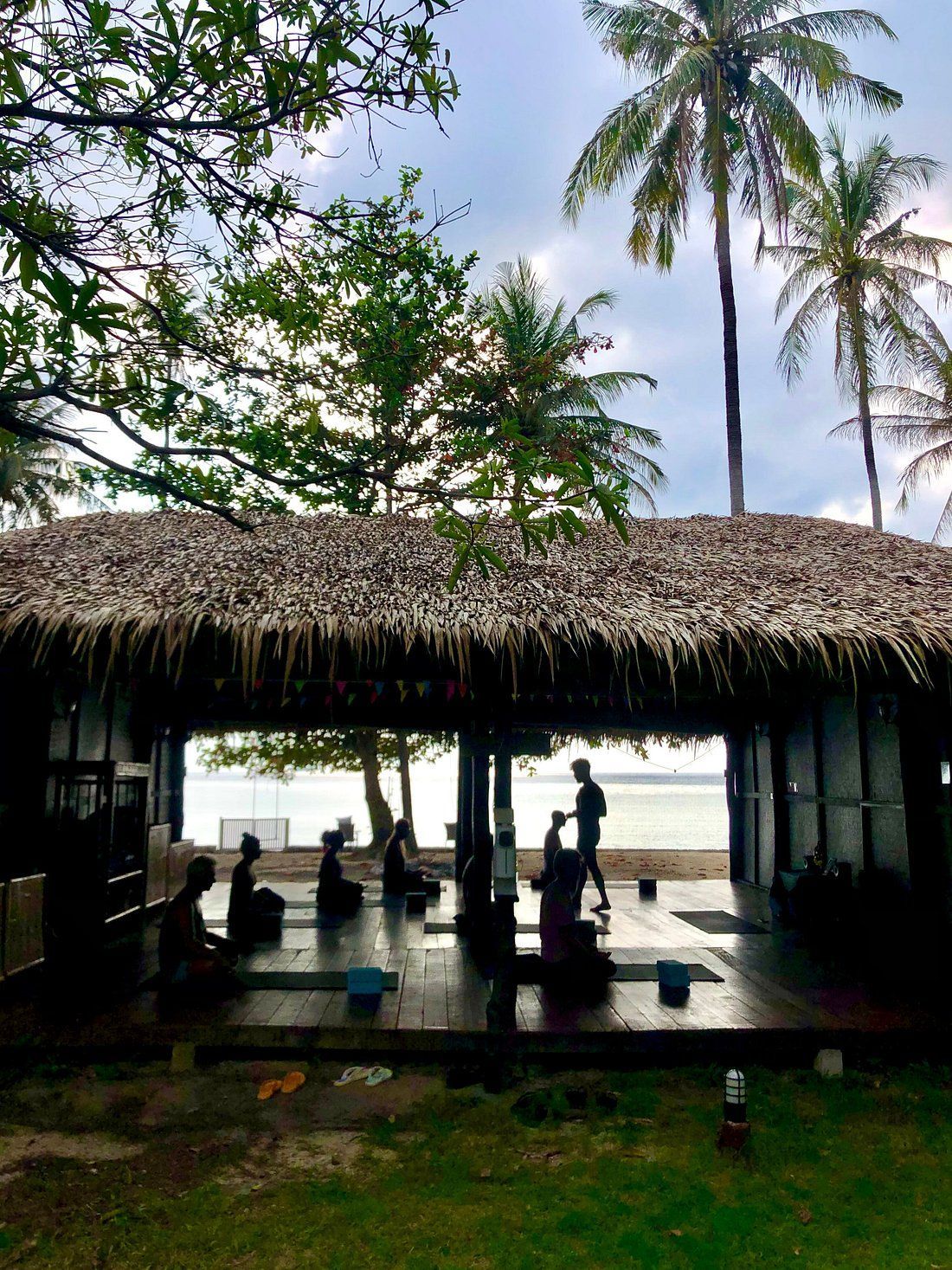 Yoga class in open-air hut by the beach. People seated, instructor standing. Palm trees, cloudy sky.