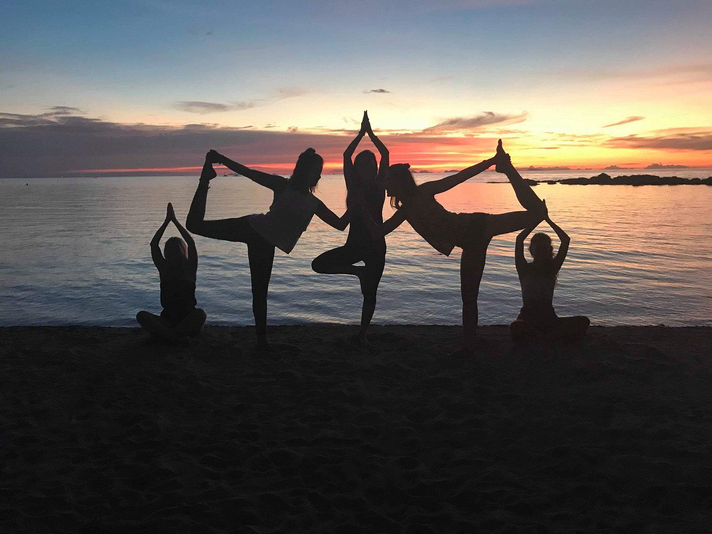 People doing yoga poses on a beach at sunset. Silhouetted figures form a star shape.