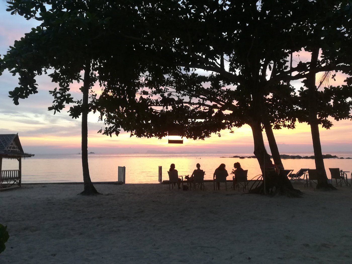 Beach at sunset with silhouetted figures seated, trees, and a gazebo.