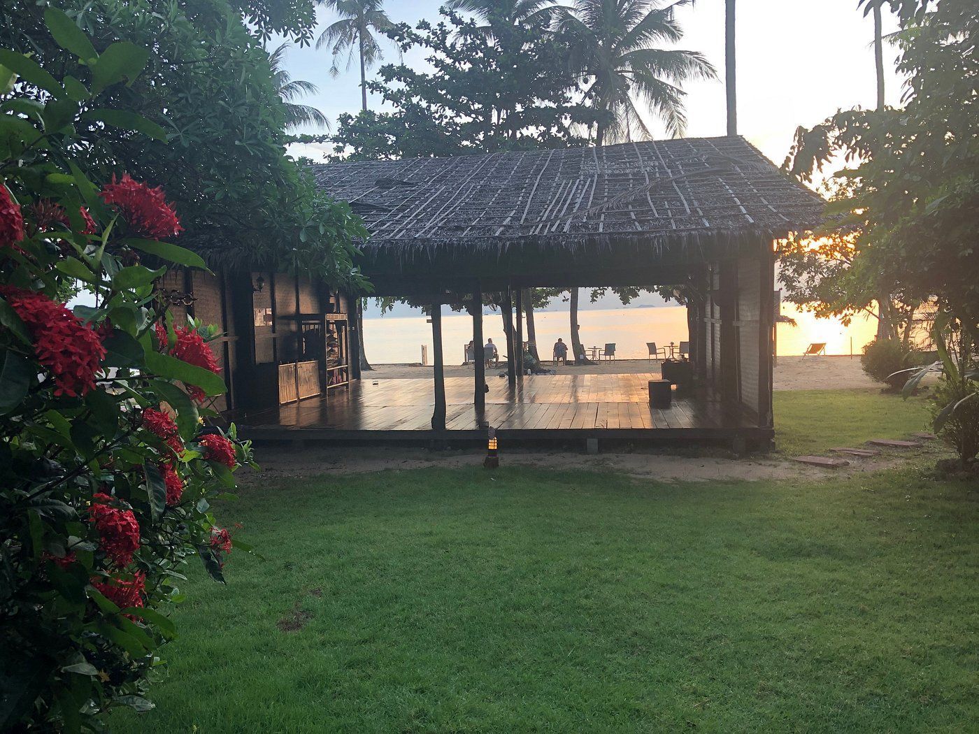 Lush green lawn with a thatched-roof structure reflecting the beach sunset. Red flowers bloom on left.