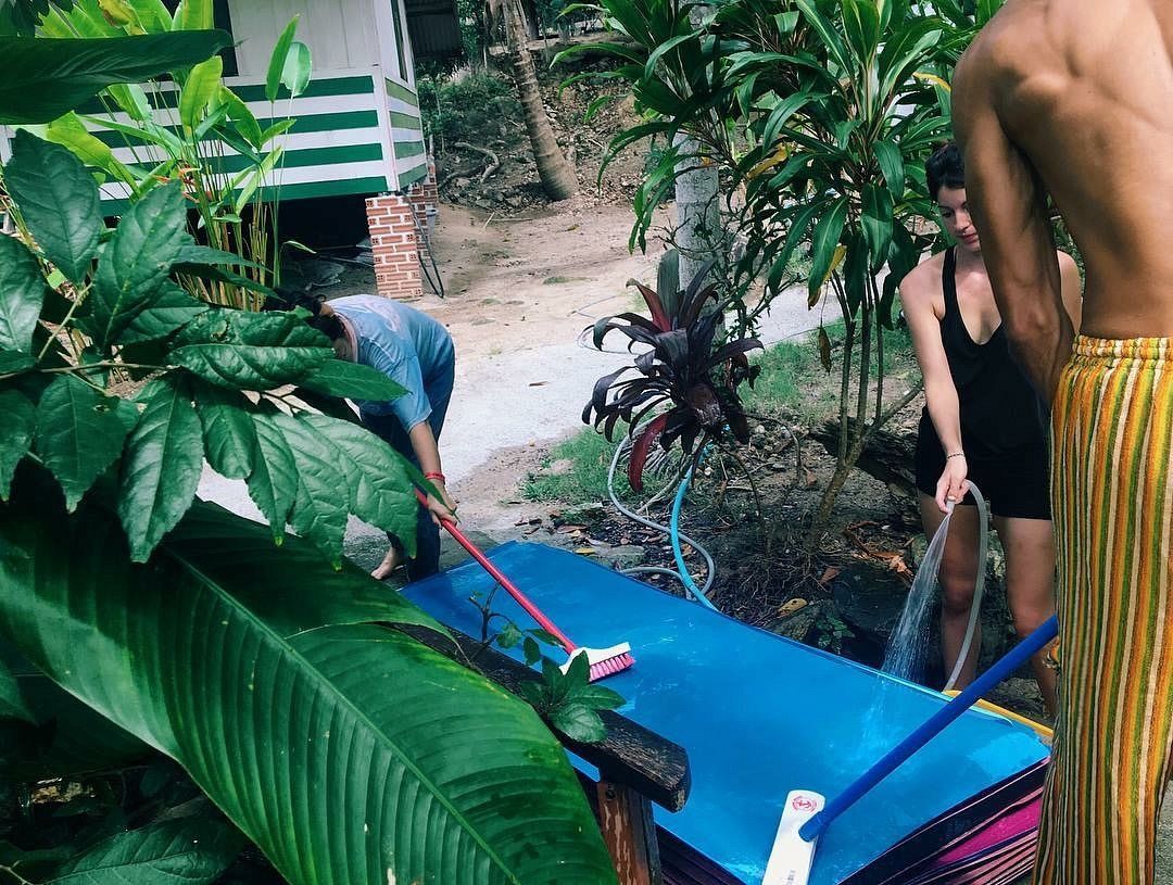 People cleaning a blue surface with mops outside, surrounded by plants and a building in the background.
