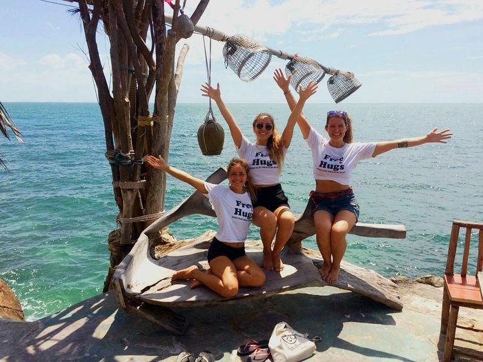 Four young women in white t-shirts with arms raised, smiling near the sea.