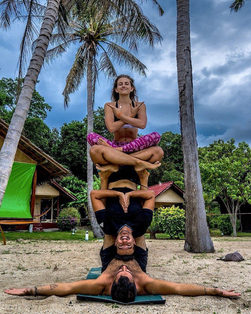 Three people in an acrobatic yoga pose on a beach with palm trees and a cloudy sky.