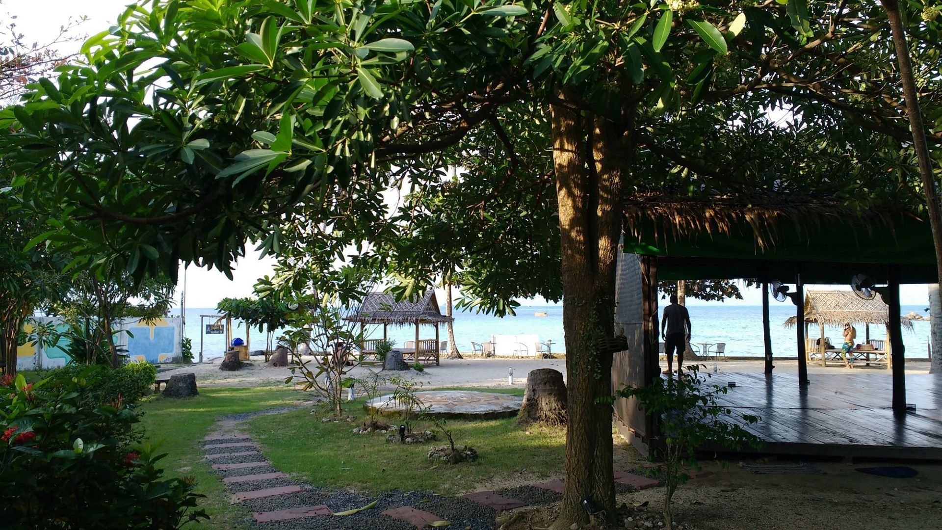 Lush garden leads to beach with gazebos. Trees provide shade; a person stands near one.