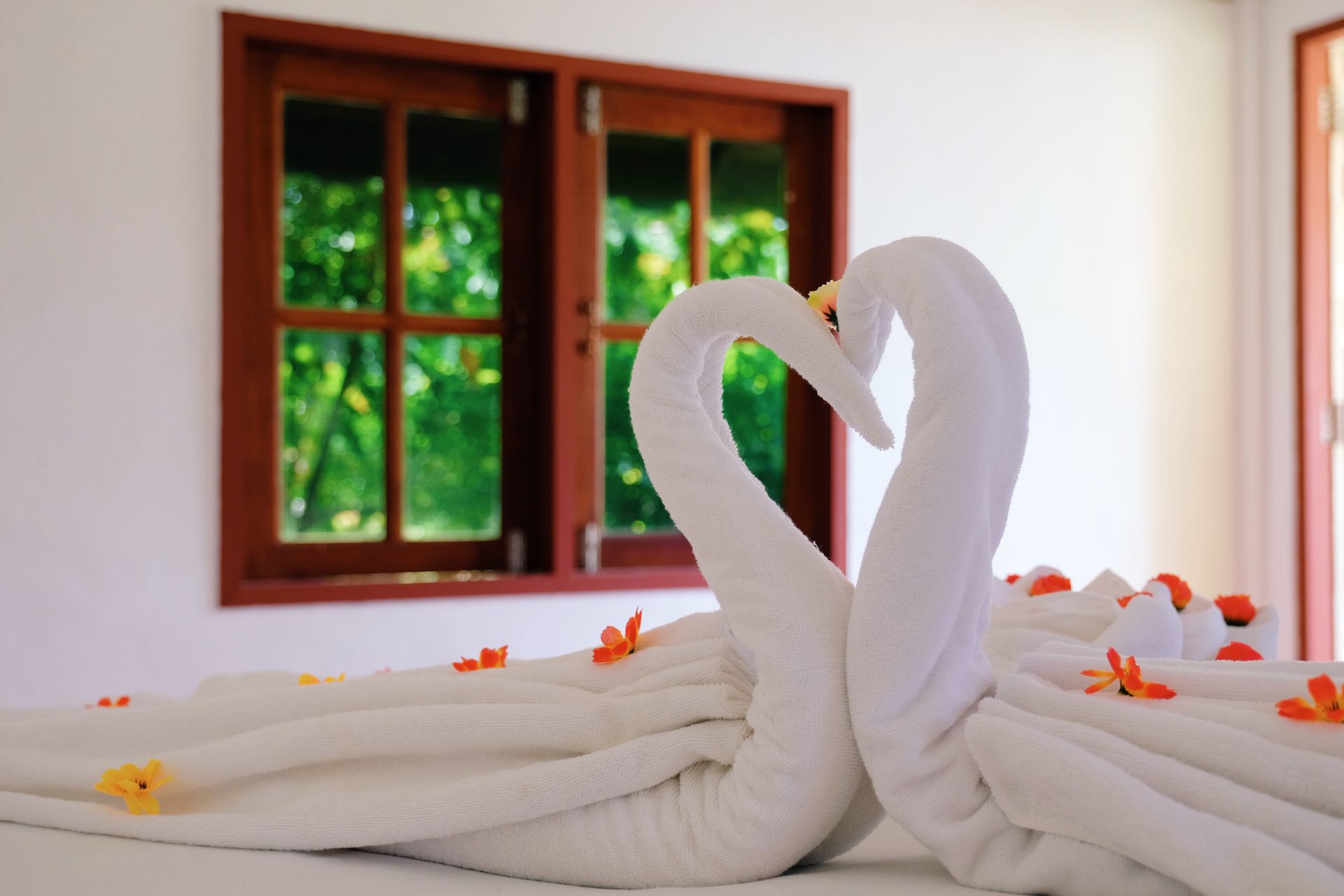 Two towel swans on a bed, forming a heart shape. Orange flowers and a window with a green view are in the background.