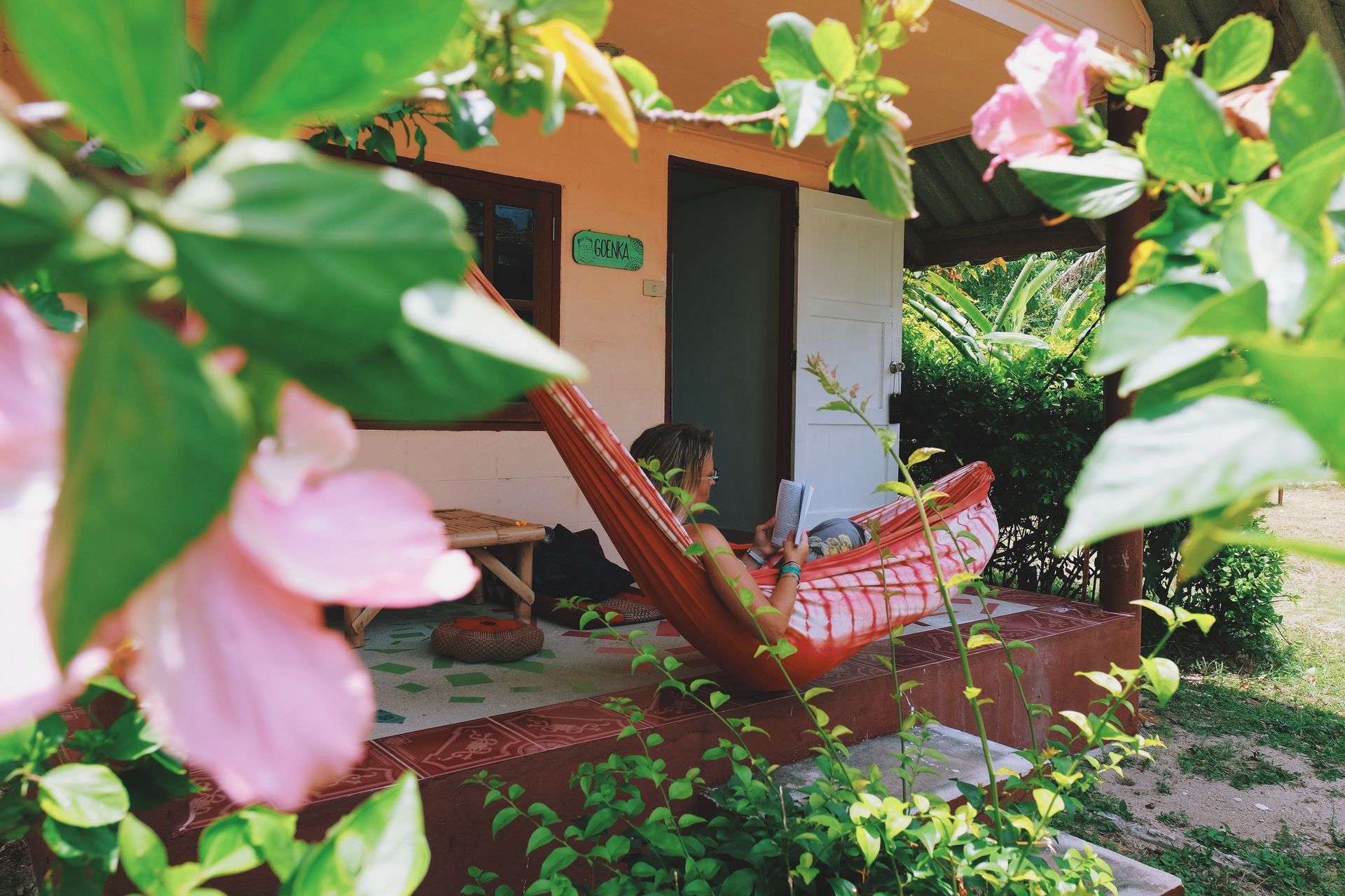 Person relaxing in a red hammock on a porch, flowers in foreground, sunny day.
