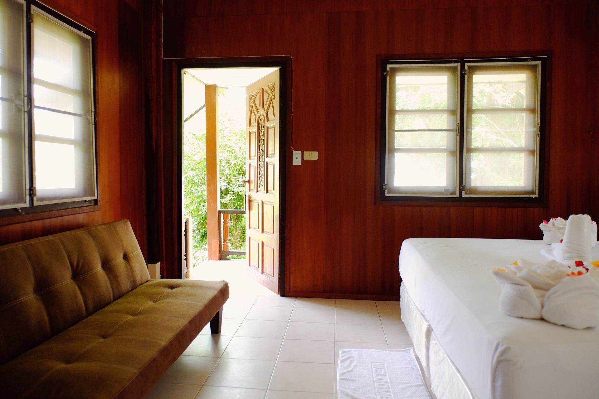 Cozy bedroom with open doorway to balcony. Brown wood paneling, sofa, bed, and sunlight.