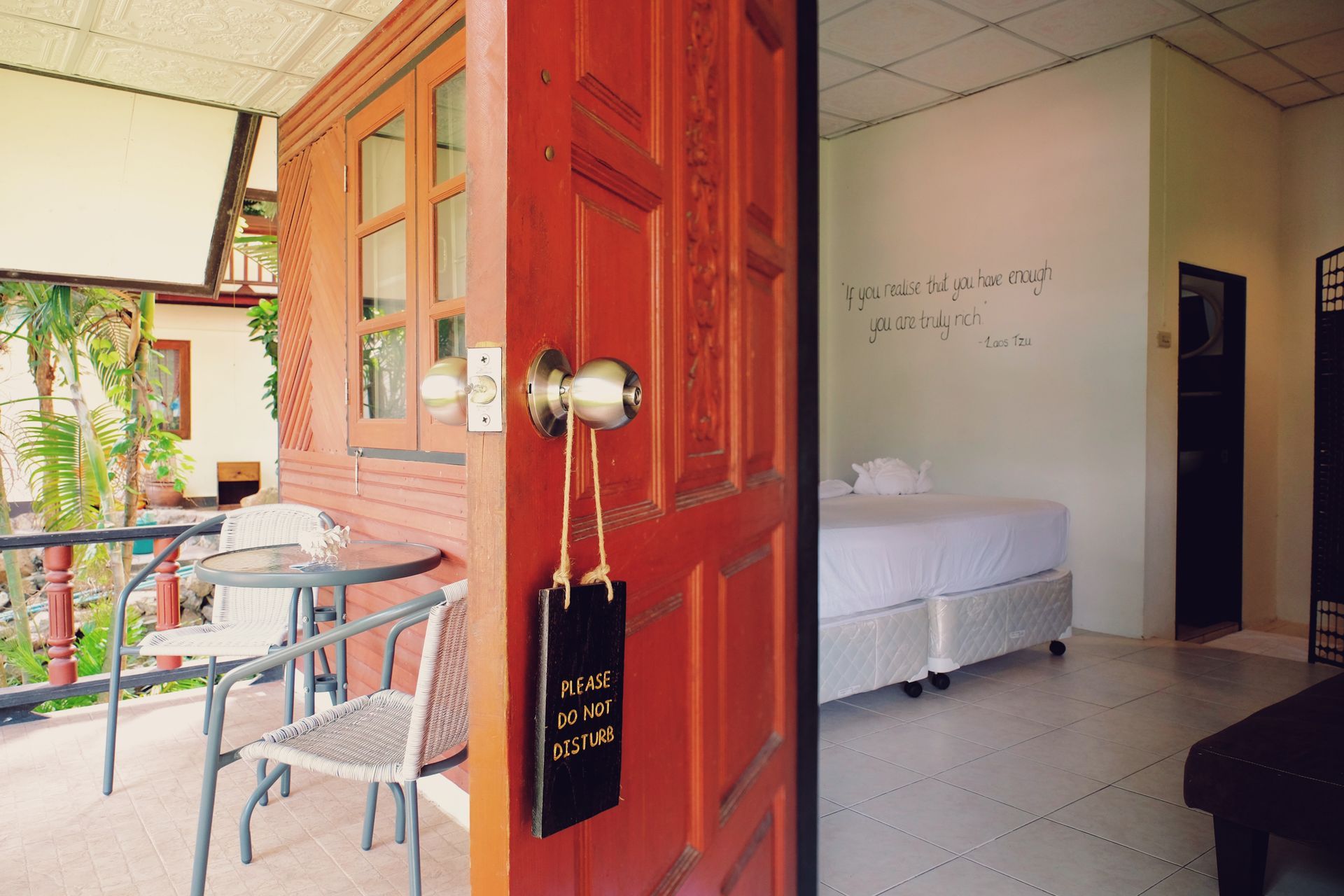 Red doorway open to a bright hotel room; a small balcony with a table and chairs on the left.