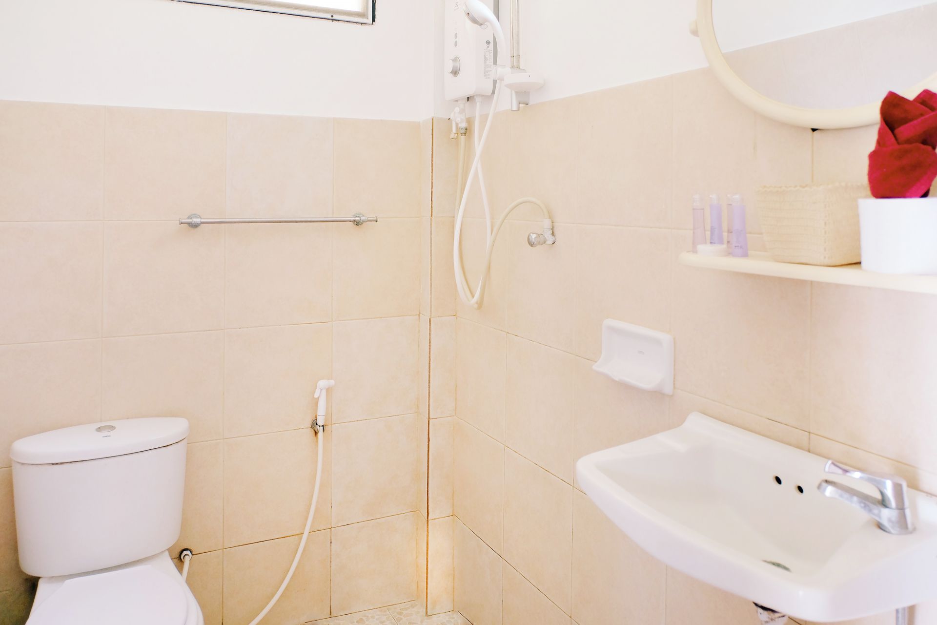 Bathroom with off-white tiled walls, a toilet, sink, and showerhead. Towel rack and shelf with toiletries are visible.