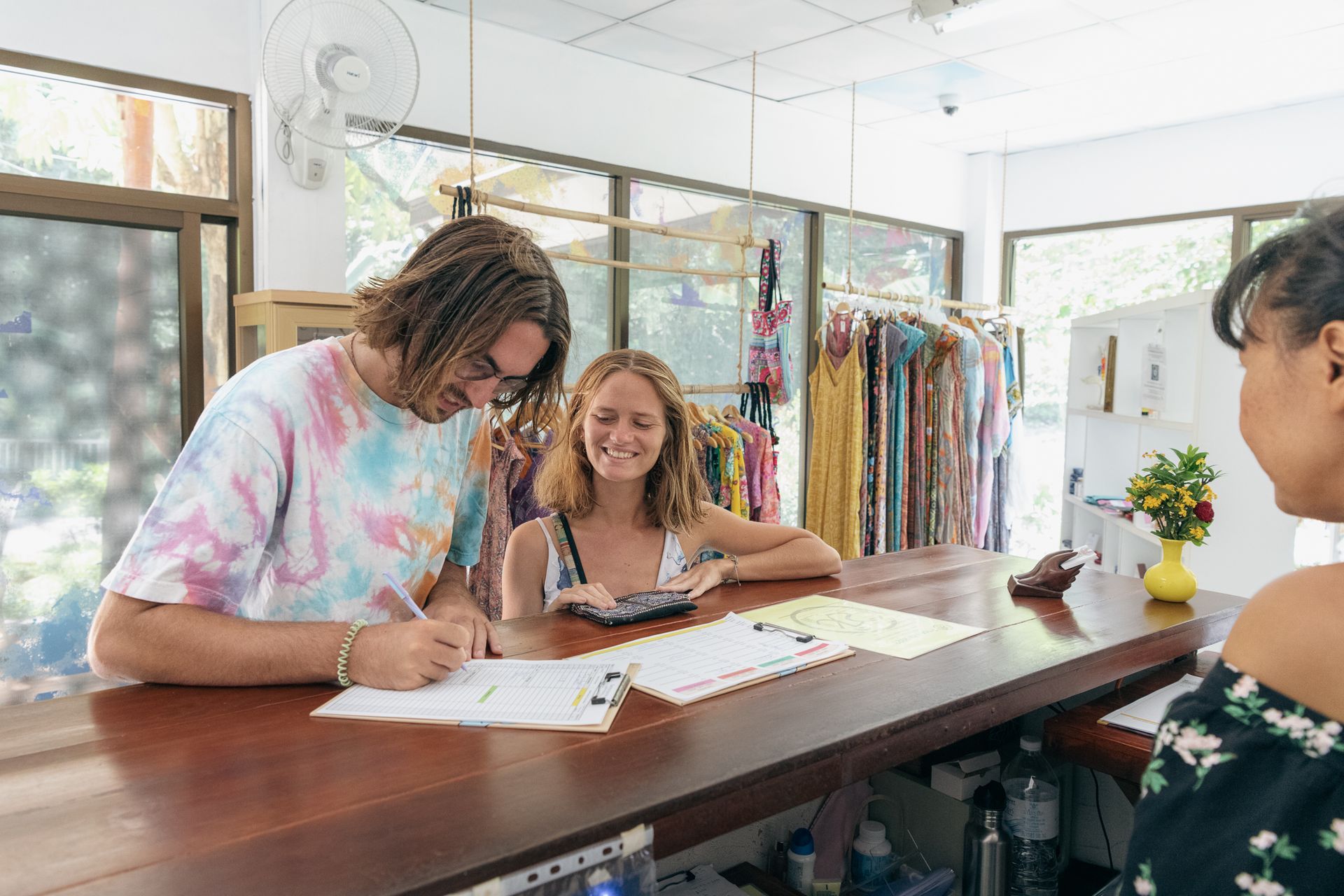 Customers at counter in a shop; man writing, woman using calculator, another woman facing them.
