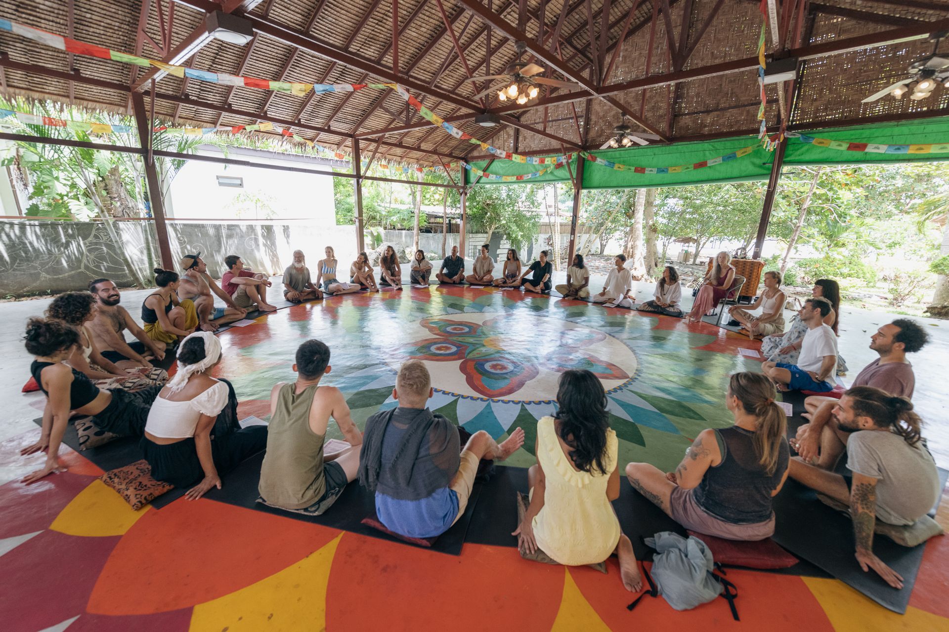 Group of people sitting in a circle on a painted floor under a round, open-air structure.