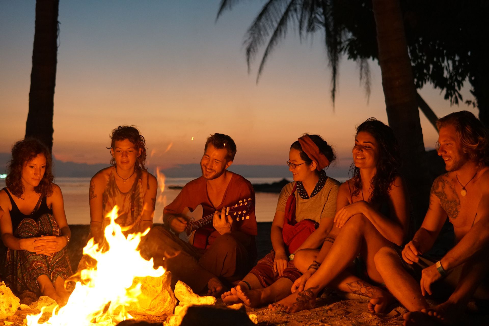 Group of friends around a bonfire on a beach at sunset, with one playing guitar.
