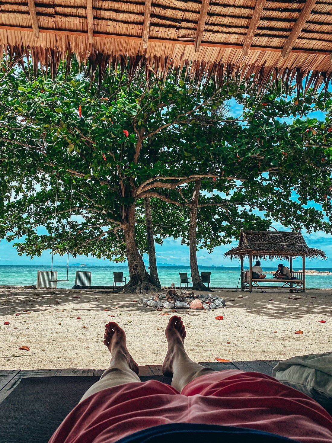 Person reclining on beach, feet forward. Sandy beach, turquoise water, trees, and thatched roof overhead.