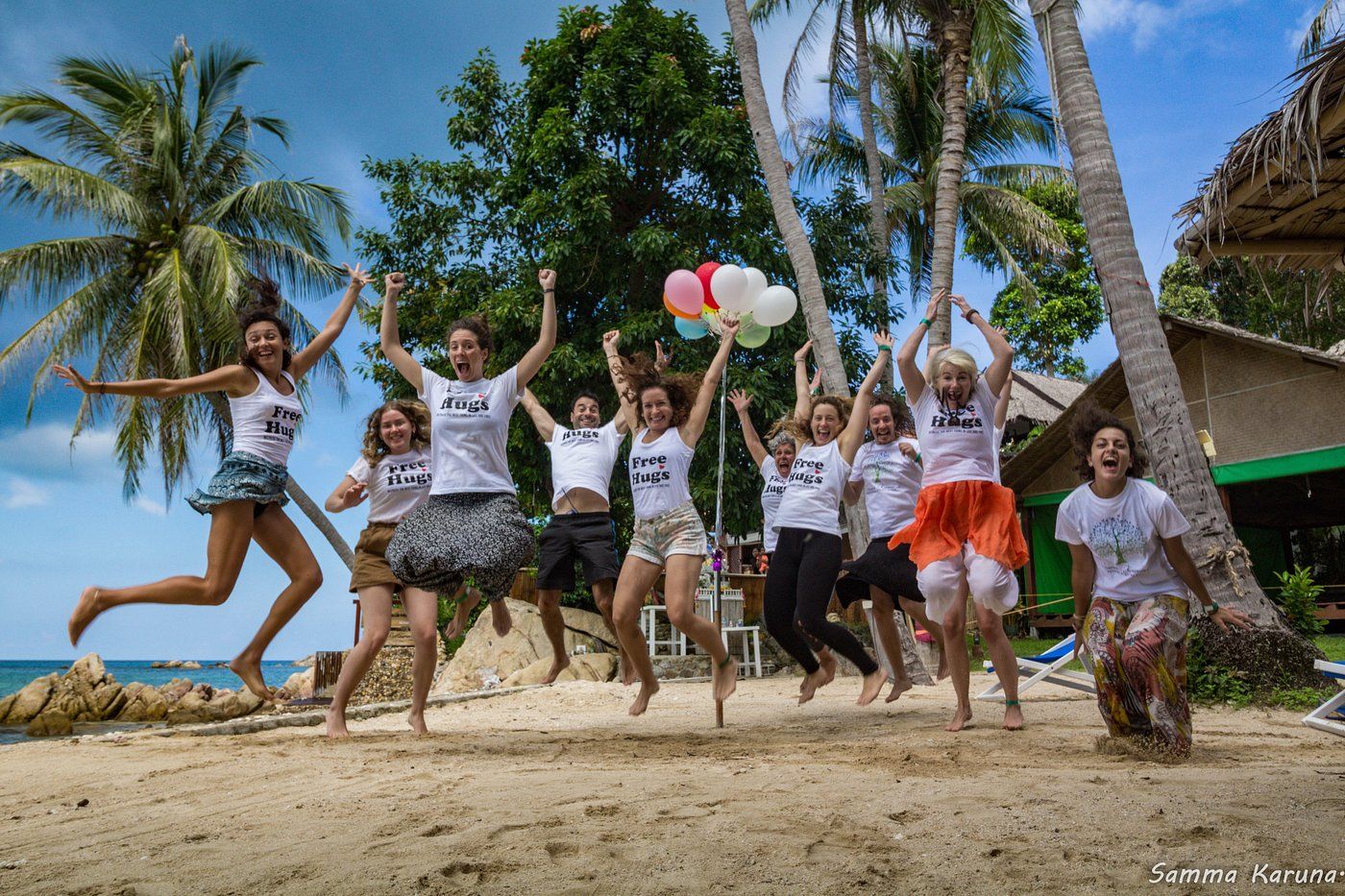 Group of young people jumping joyfully on a tropical beach, white shirts, colorful skirts, balloons.