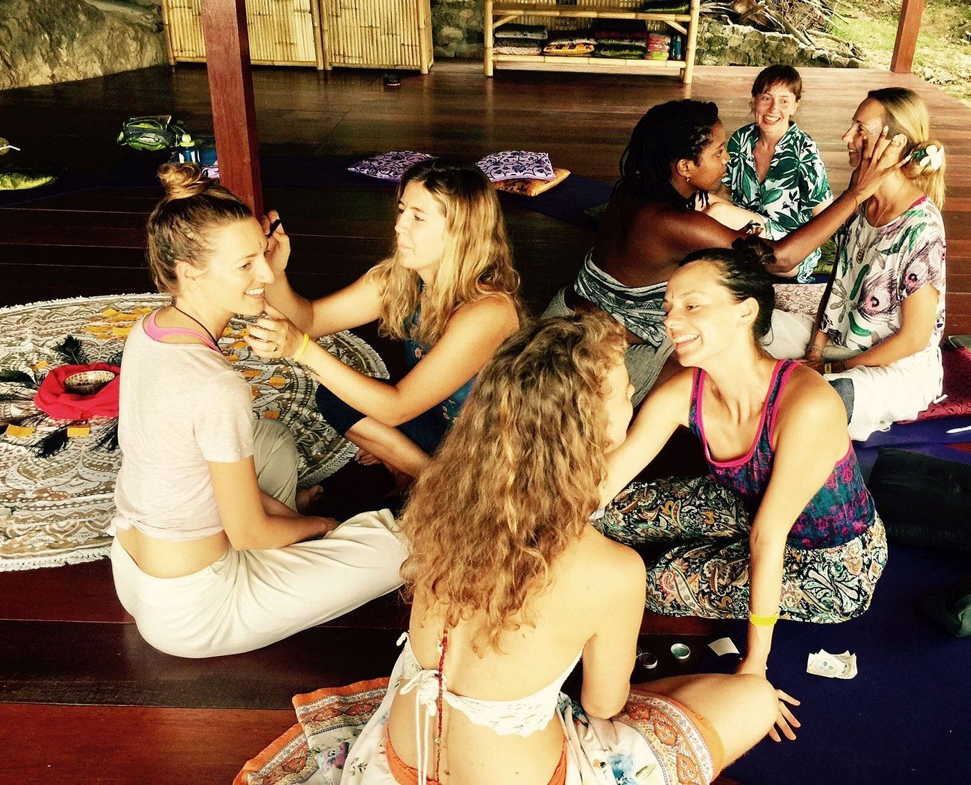 Group of women in a circle, hands touching faces. Smiling, diverse skin tones, outdoor wooden setting.