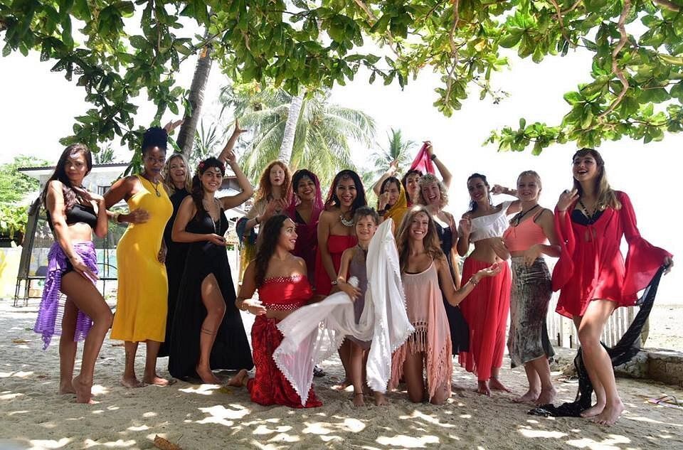 Group of women in colorful dresses posing on a beach under a tree.